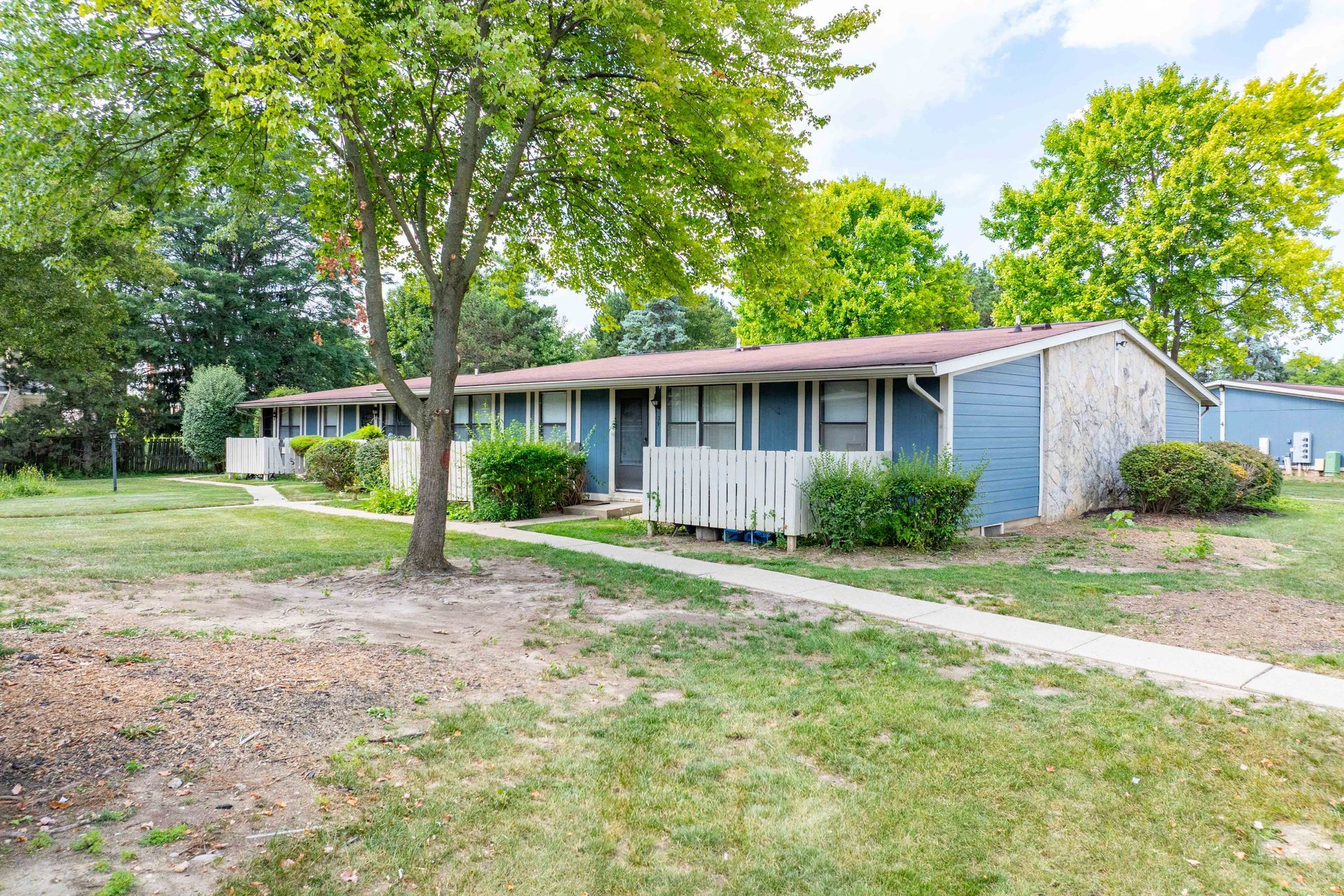 Low-slung blue and brick-colored buildings with white fences, a tree, and grassy yard.