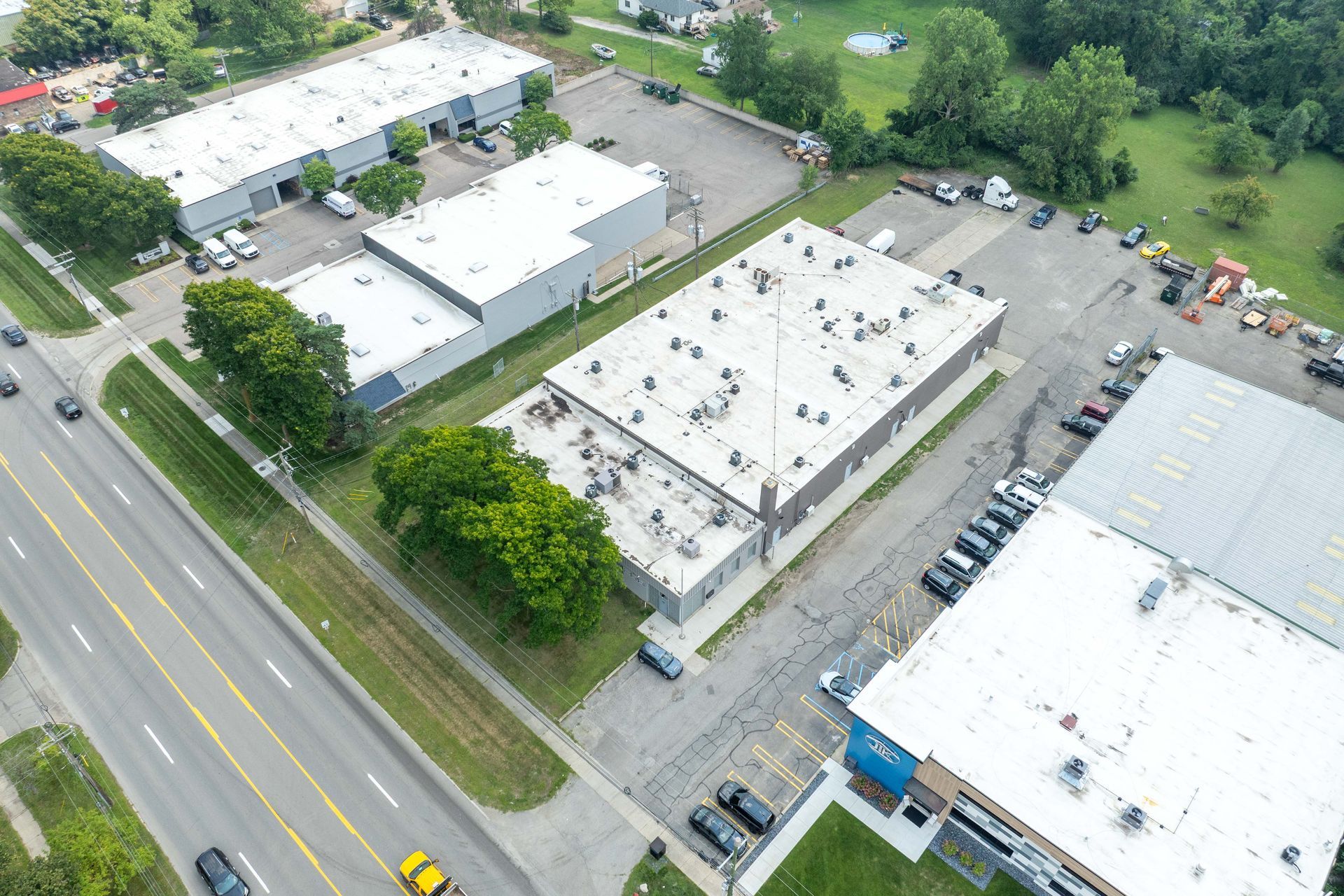 Aerial view of industrial buildings with parking lots, surrounded by trees and a road with traffic.