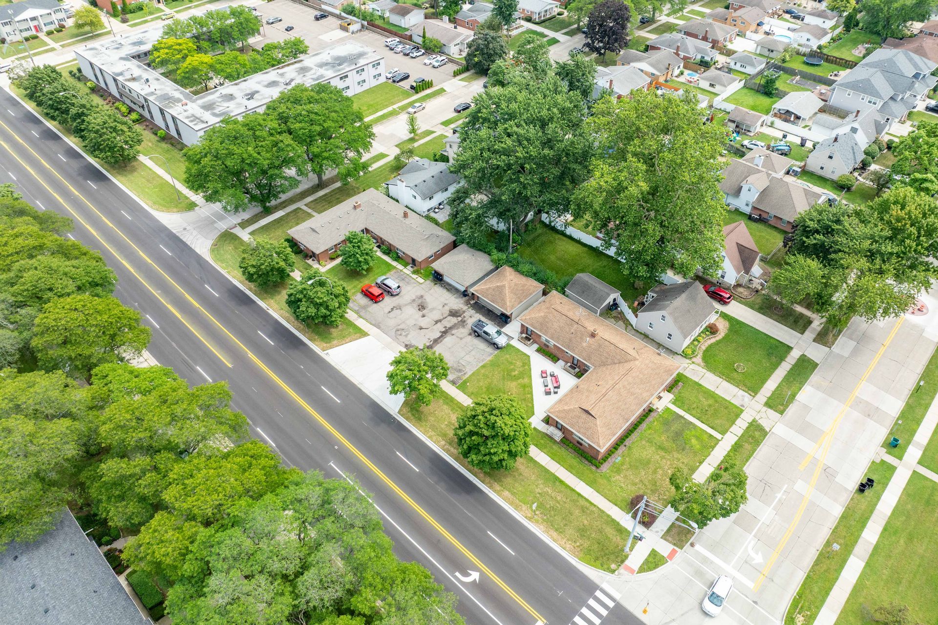 Aerial view of houses and a road with trees. Green grass and blue sky are visible.