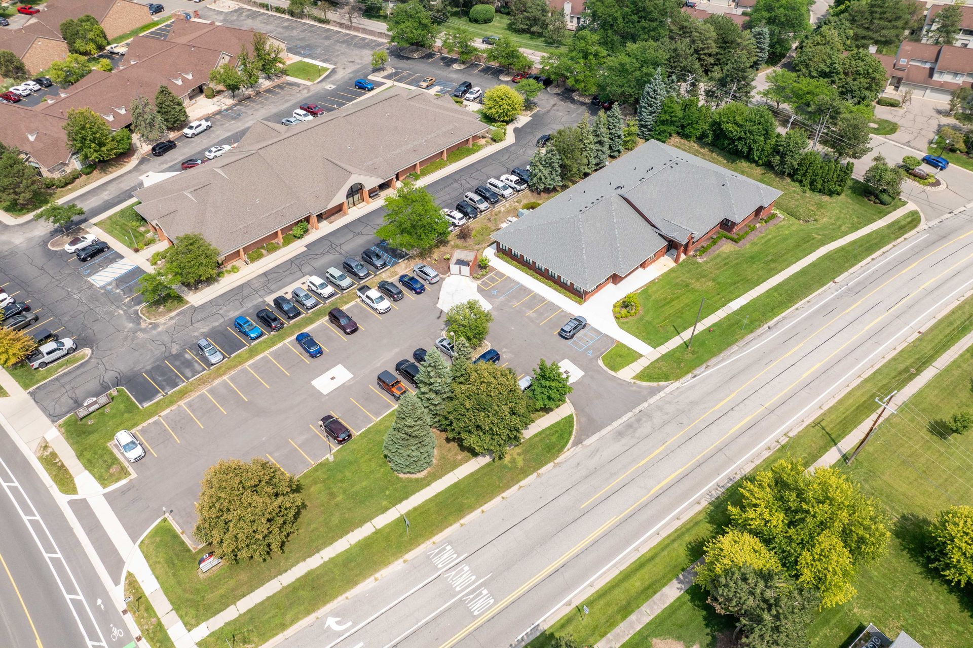 Aerial view of two brown-roofed buildings with parking, trees, and a road on a sunny day.