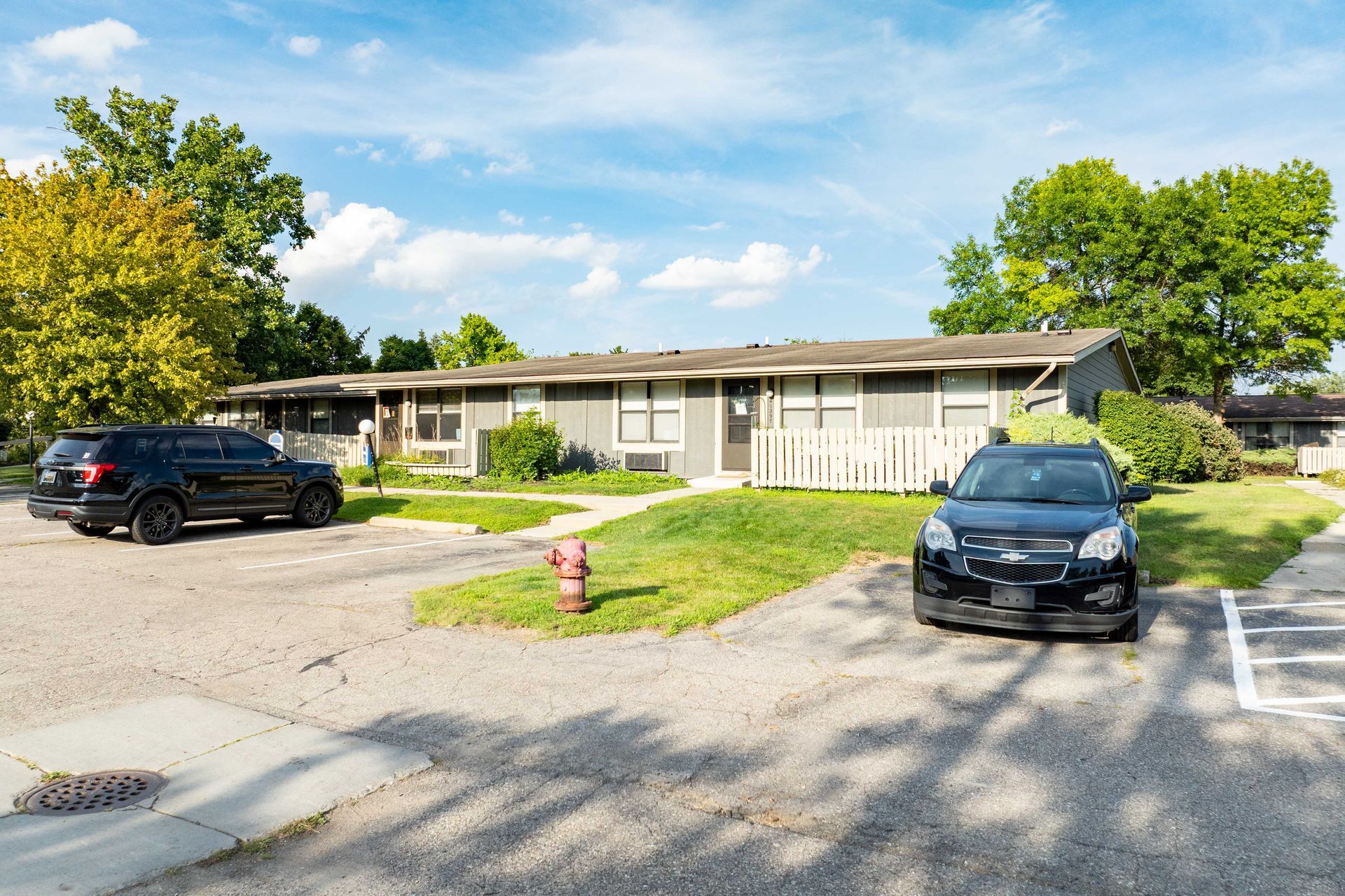 A gray, one-story building with a low-sloping roof, with two cars parked in front of it on a sunny day.