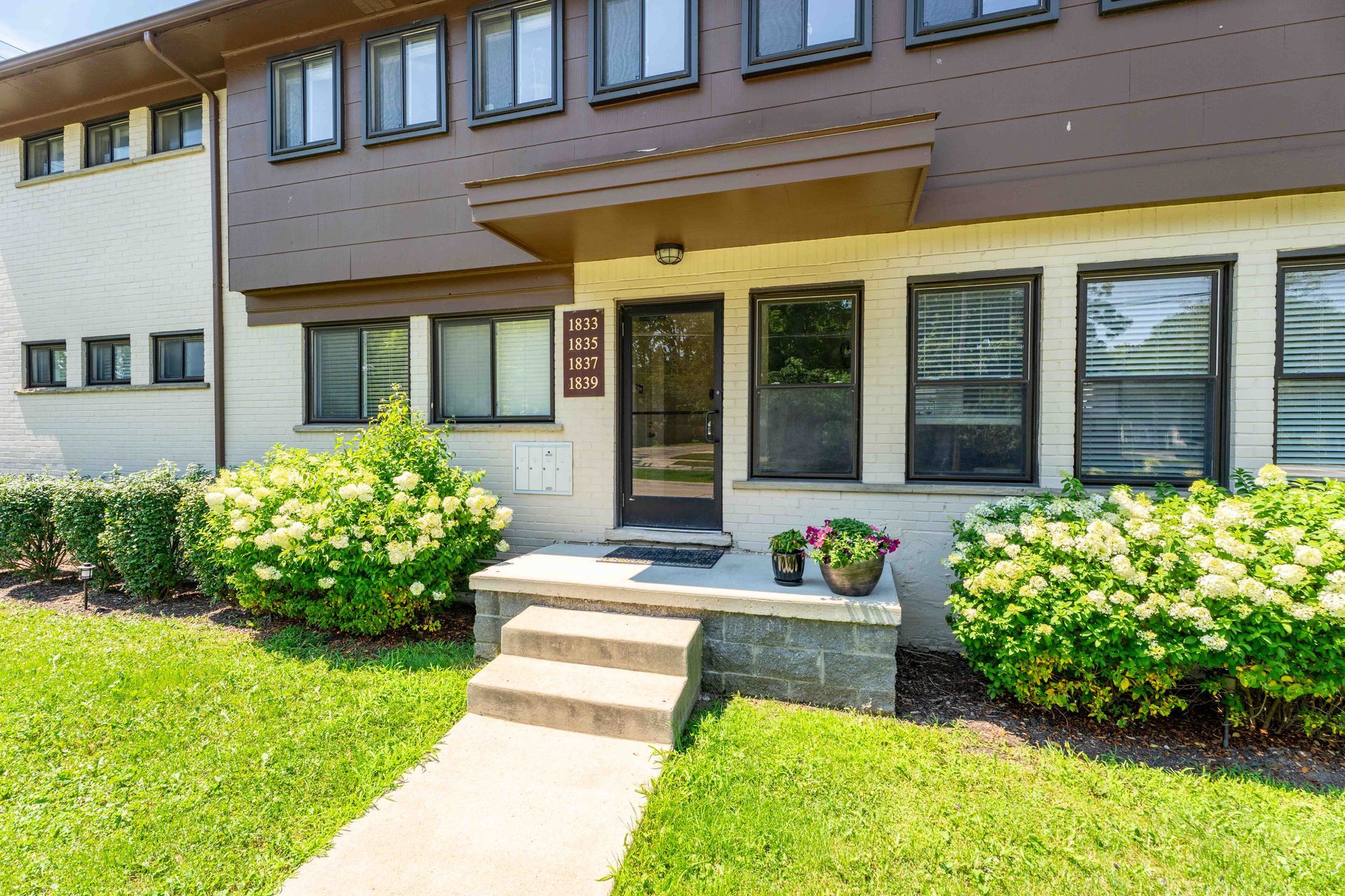 Brown and white townhouse exterior with front steps, shrubs, and green lawn.