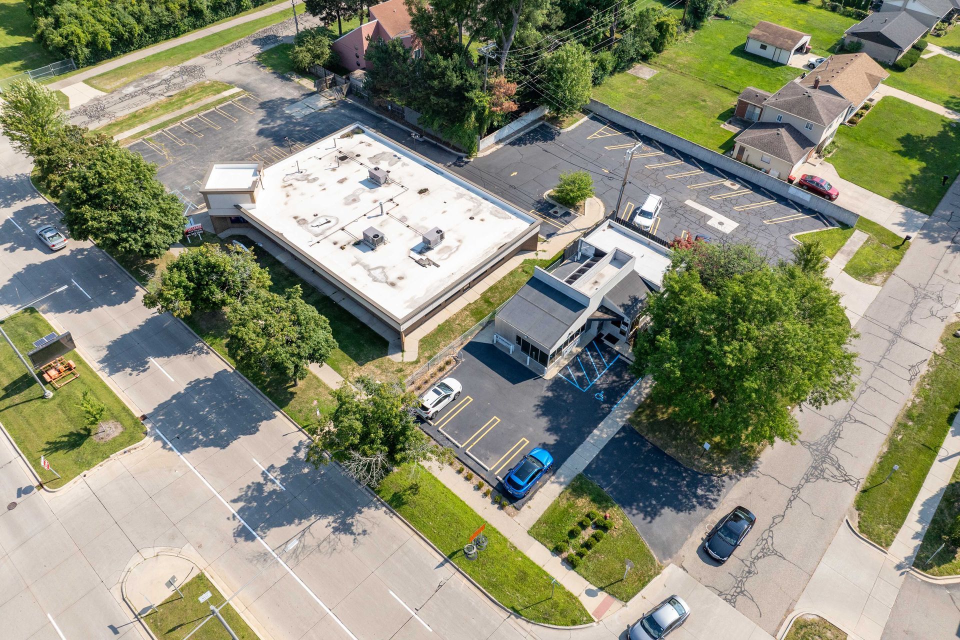 Aerial view of commercial buildings, parking lots, and a street surrounded by trees and green lawns.