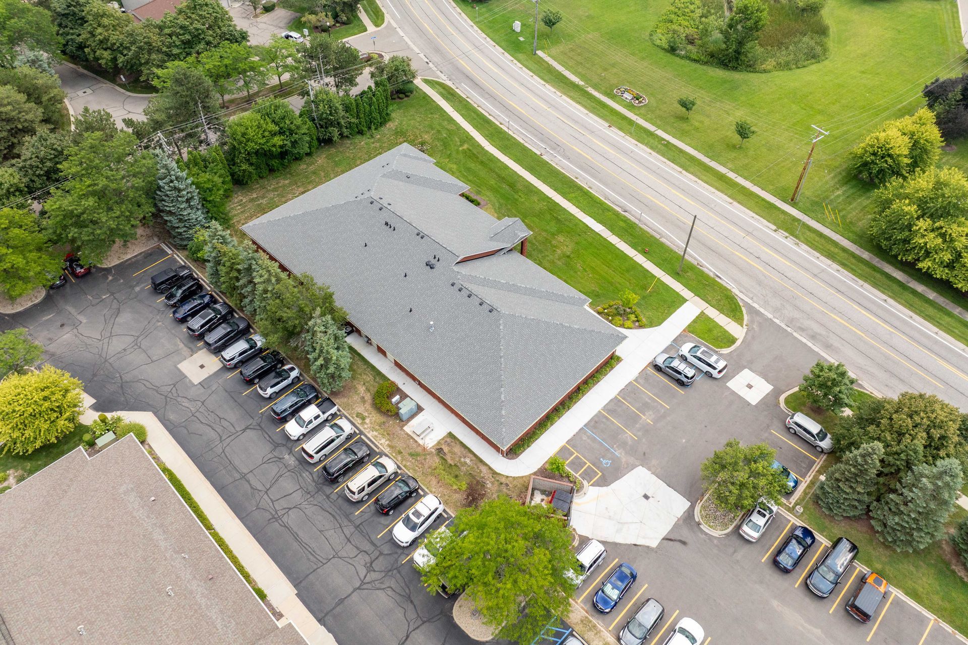 Aerial view of a building with a gray roof and parking lots on either side; a road is visible in the background.