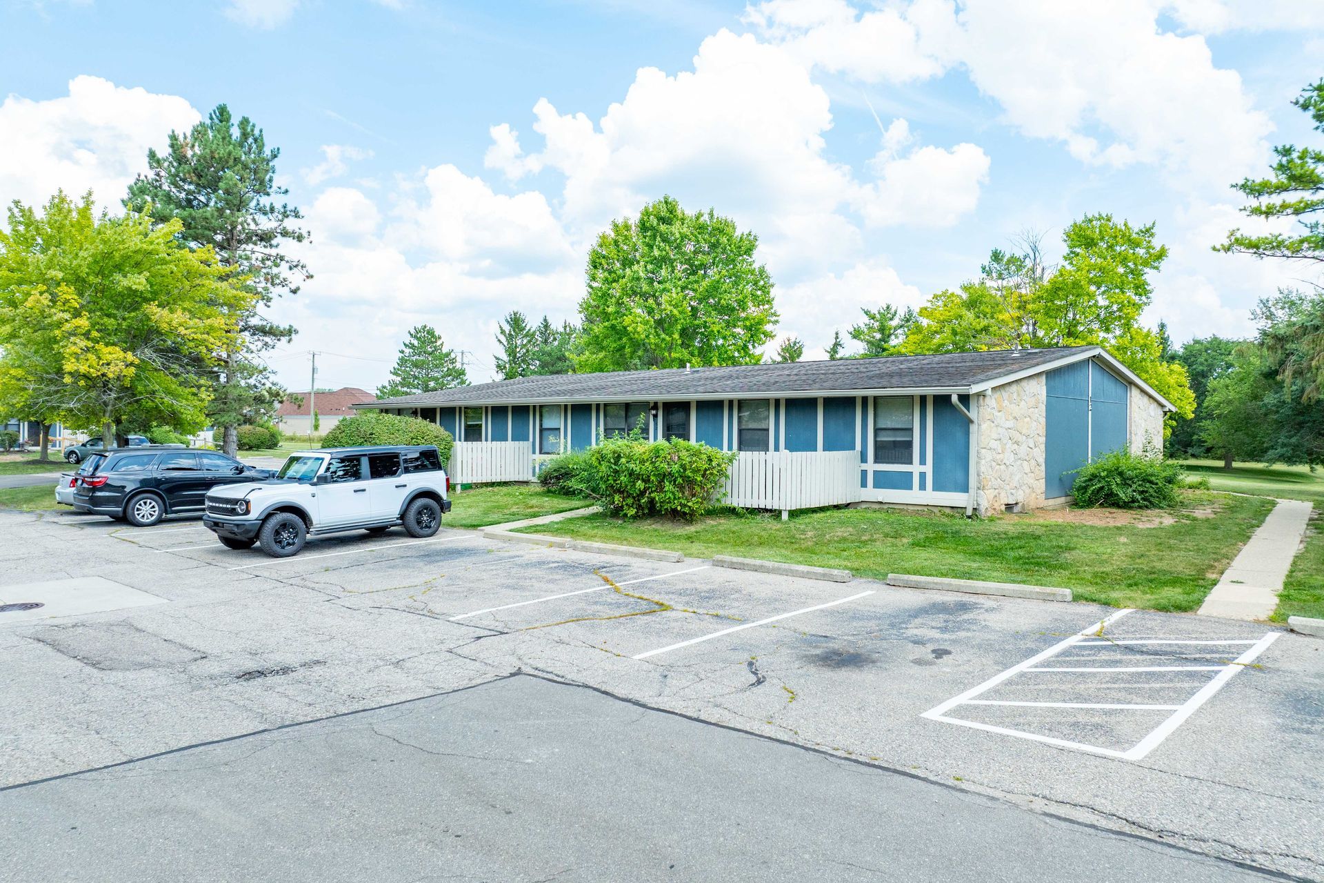 A blue and brick building with parking lot. White SUV parked in front. Sunny day, some trees.