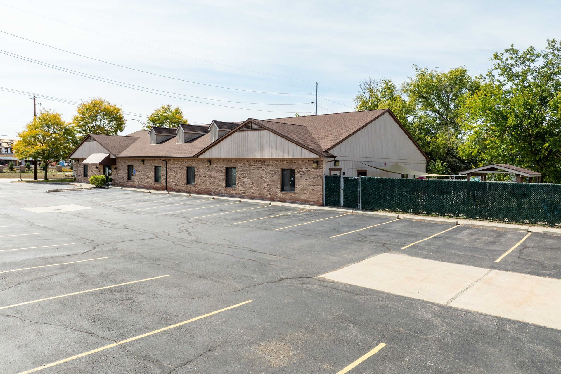 An empty parking lot in front of a low-rise brick building with a brown roof and trees on a sunny day.