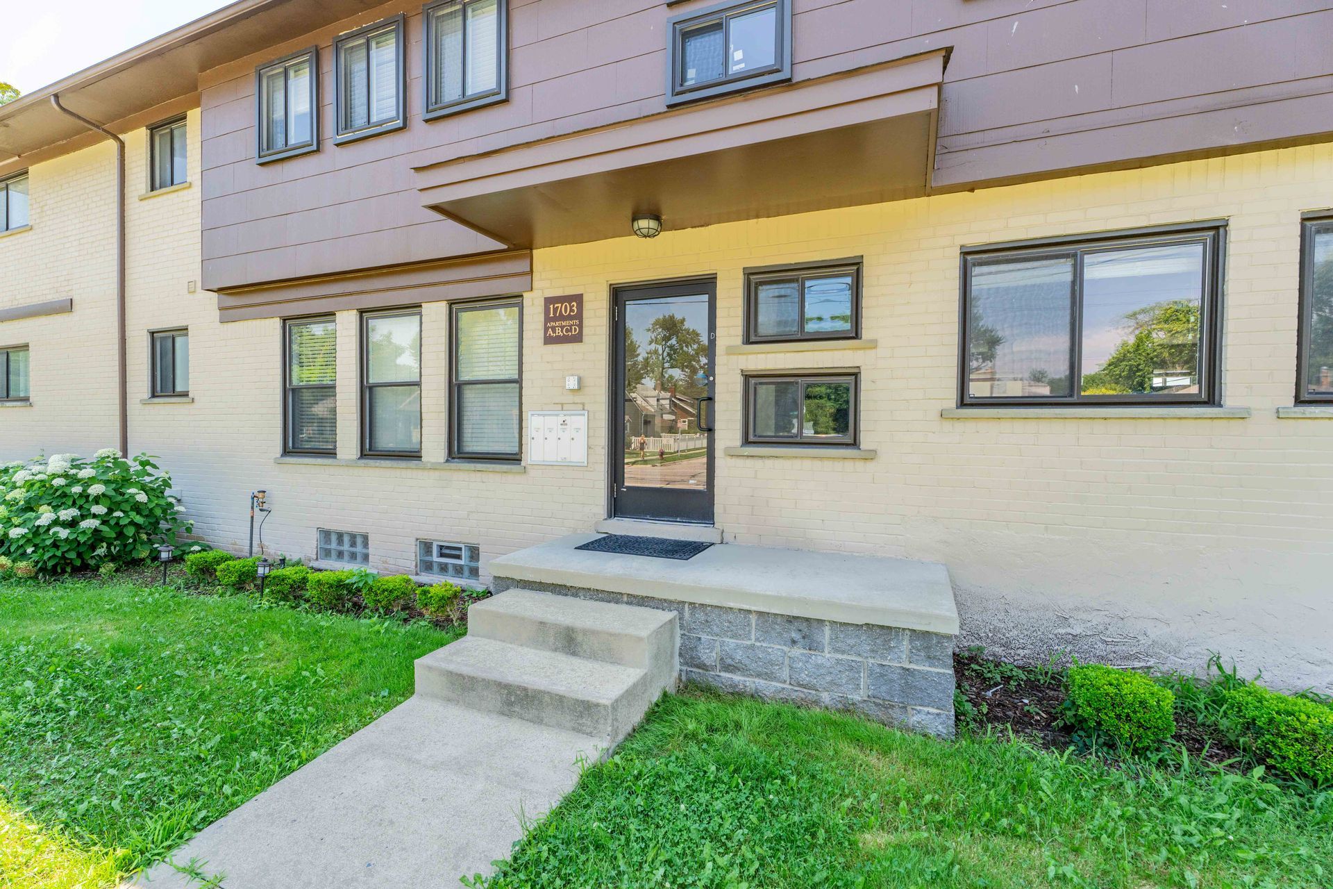 Tan brick apartment building with a concrete stoop and small front yard.