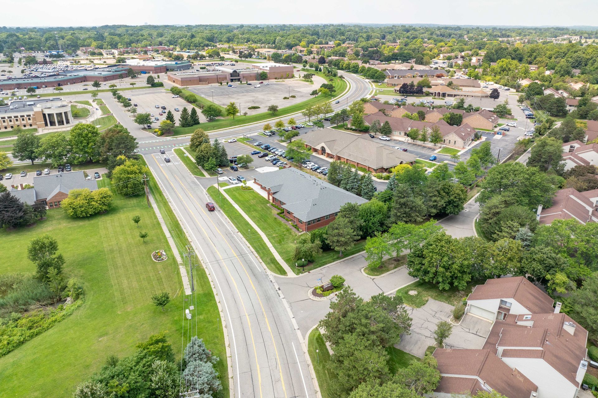 Aerial view of a suburban road, surrounded by buildings, trees, and green lawns, on a bright day.