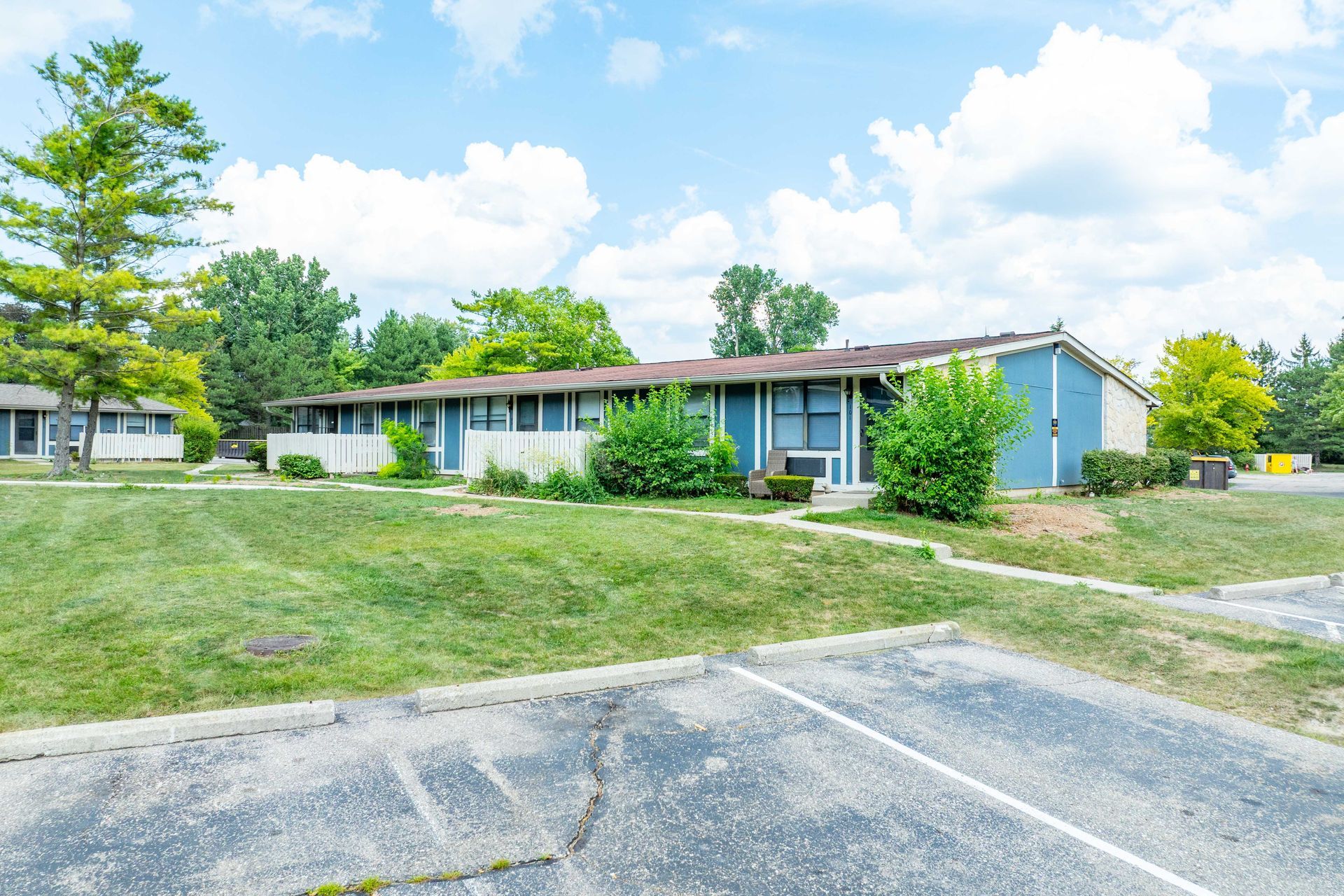 Blue apartment buildings with green bushes and a parking lot under a blue sky.