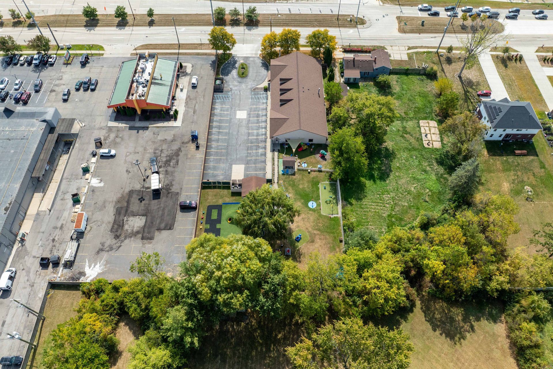 Aerial view of a commercial area with a parking lot, buildings, trees, and a road.