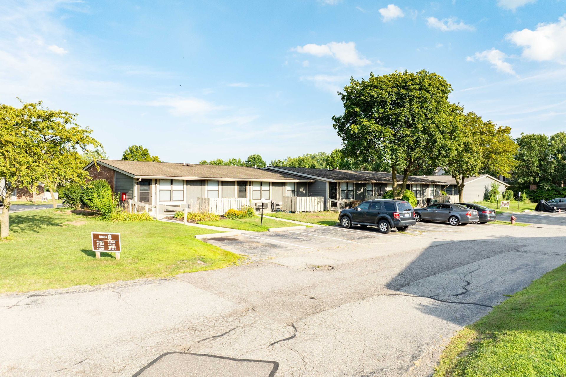 Low-rise apartment buildings with cars parked in front on a sunny day. Green grass and trees surround.