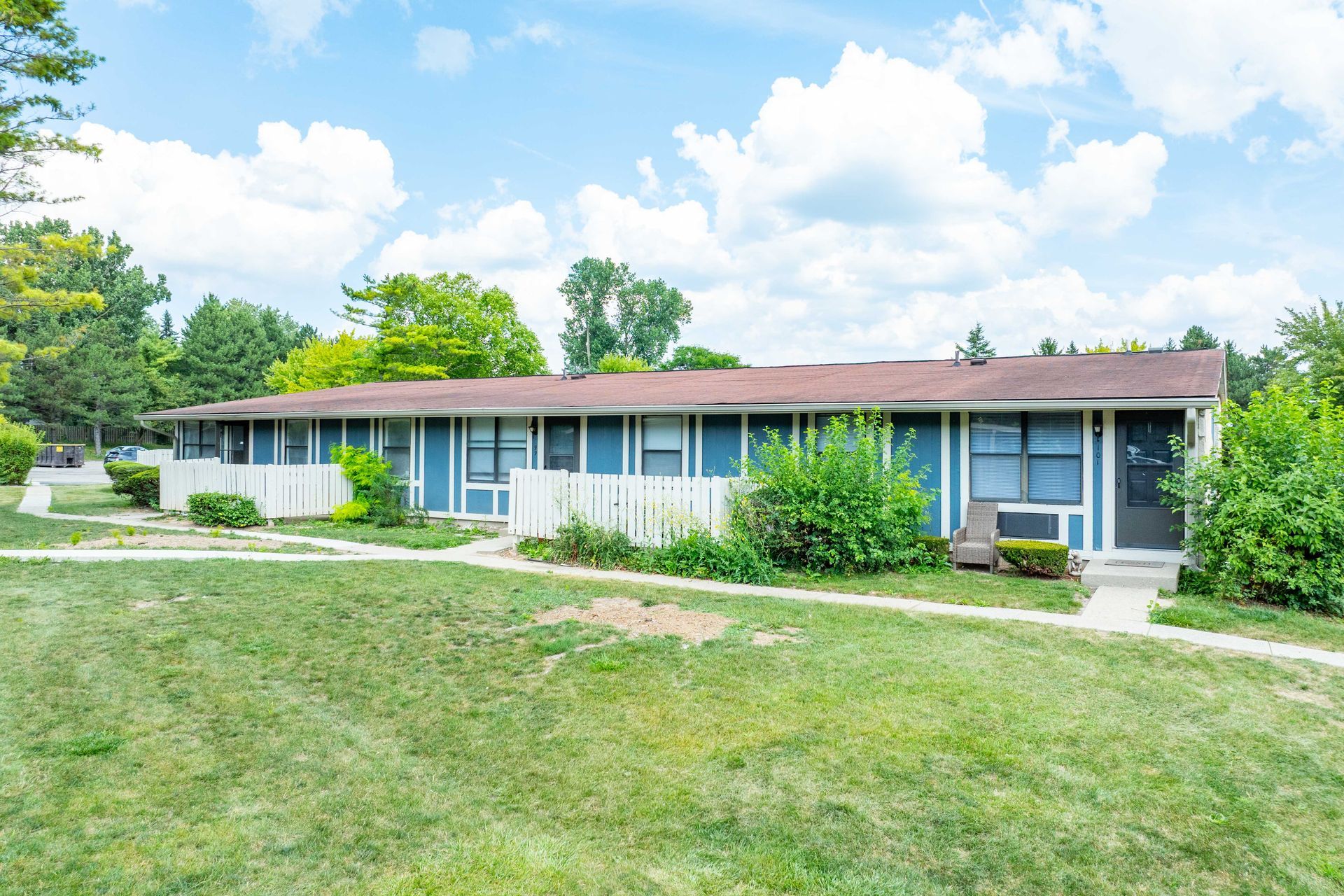Blue apartment building with white fence and green lawn under a cloudy sky.