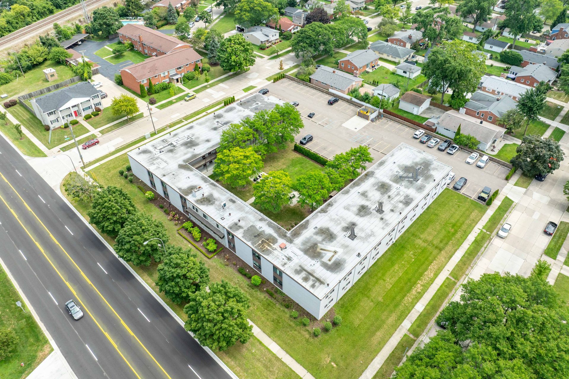 Aerial view of a white rectangular apartment complex with a central green courtyard surrounded by trees and a street with lanes.