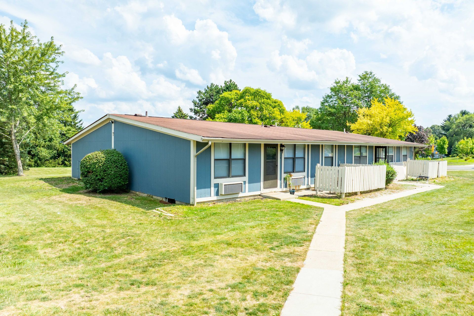 Blue apartment building with brown roof, fronted by a grassy lawn and a sidewalk on a sunny day.