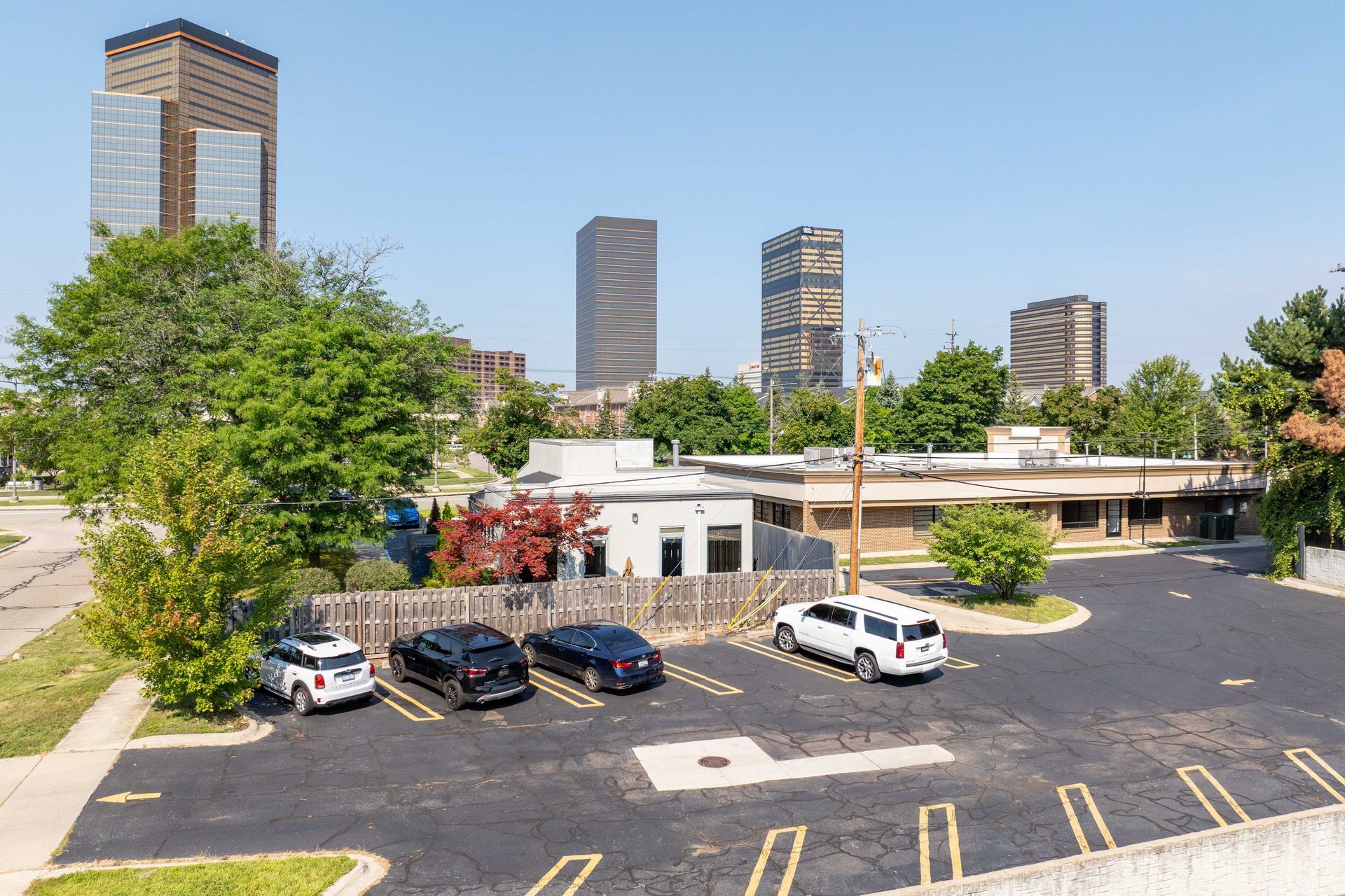 Vehicles parked in front of a building with tall skyscrapers in the background on a sunny day.