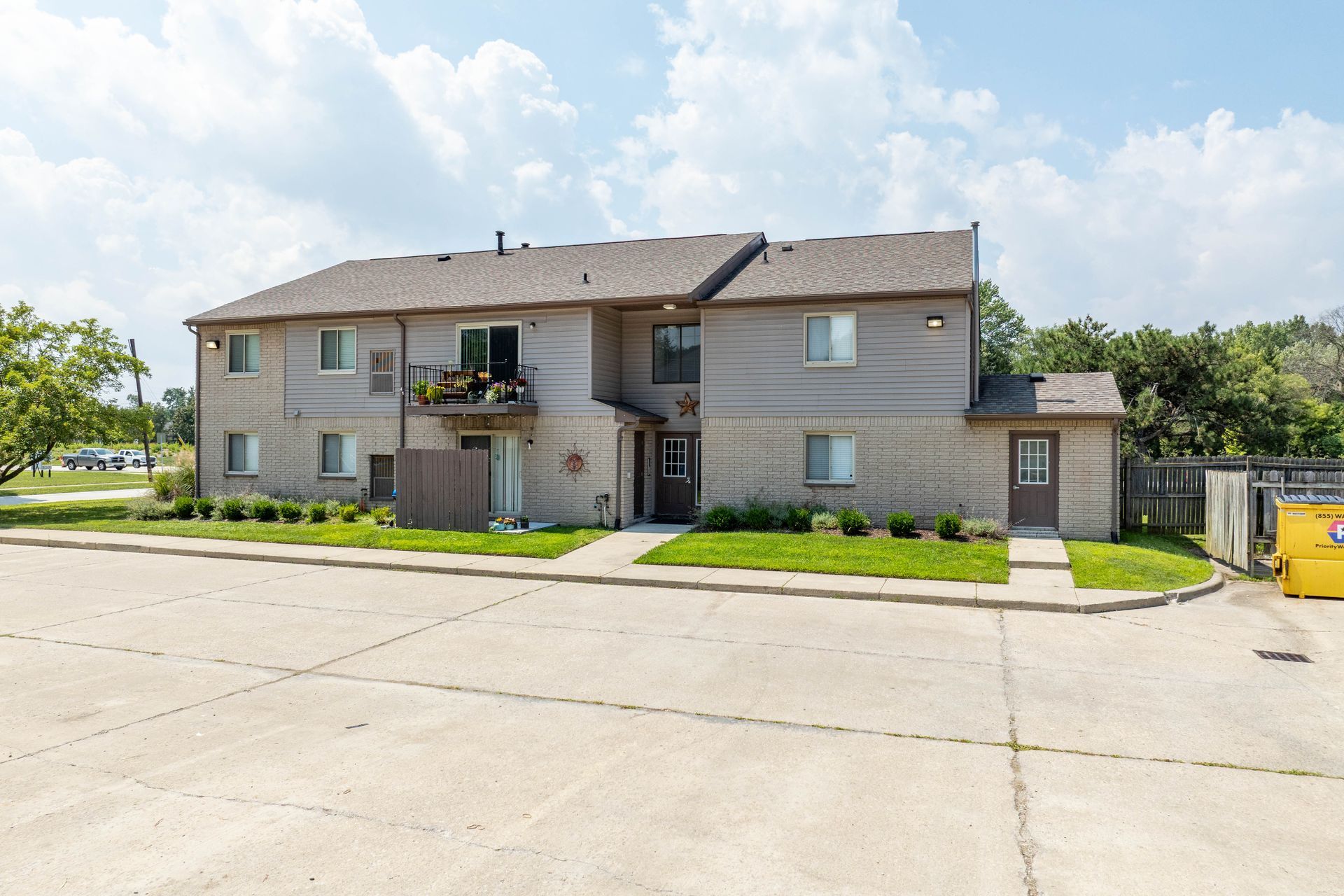 Two-story brick apartment building with tan roof, gray walls, and small balcony. Sunny day.