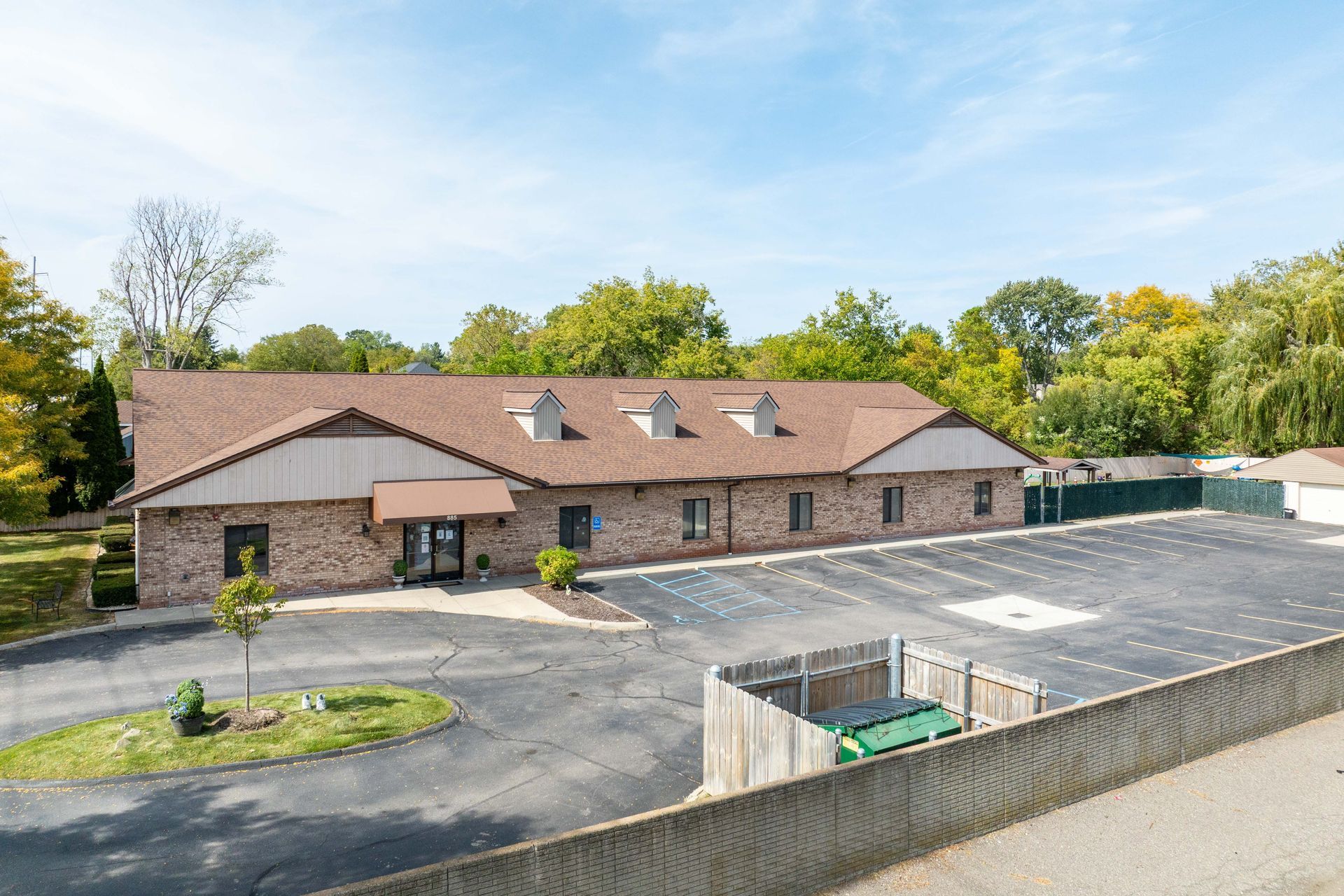 Brick building with brown roof, dormers, and large parking lot. Trees in background, sunny day.