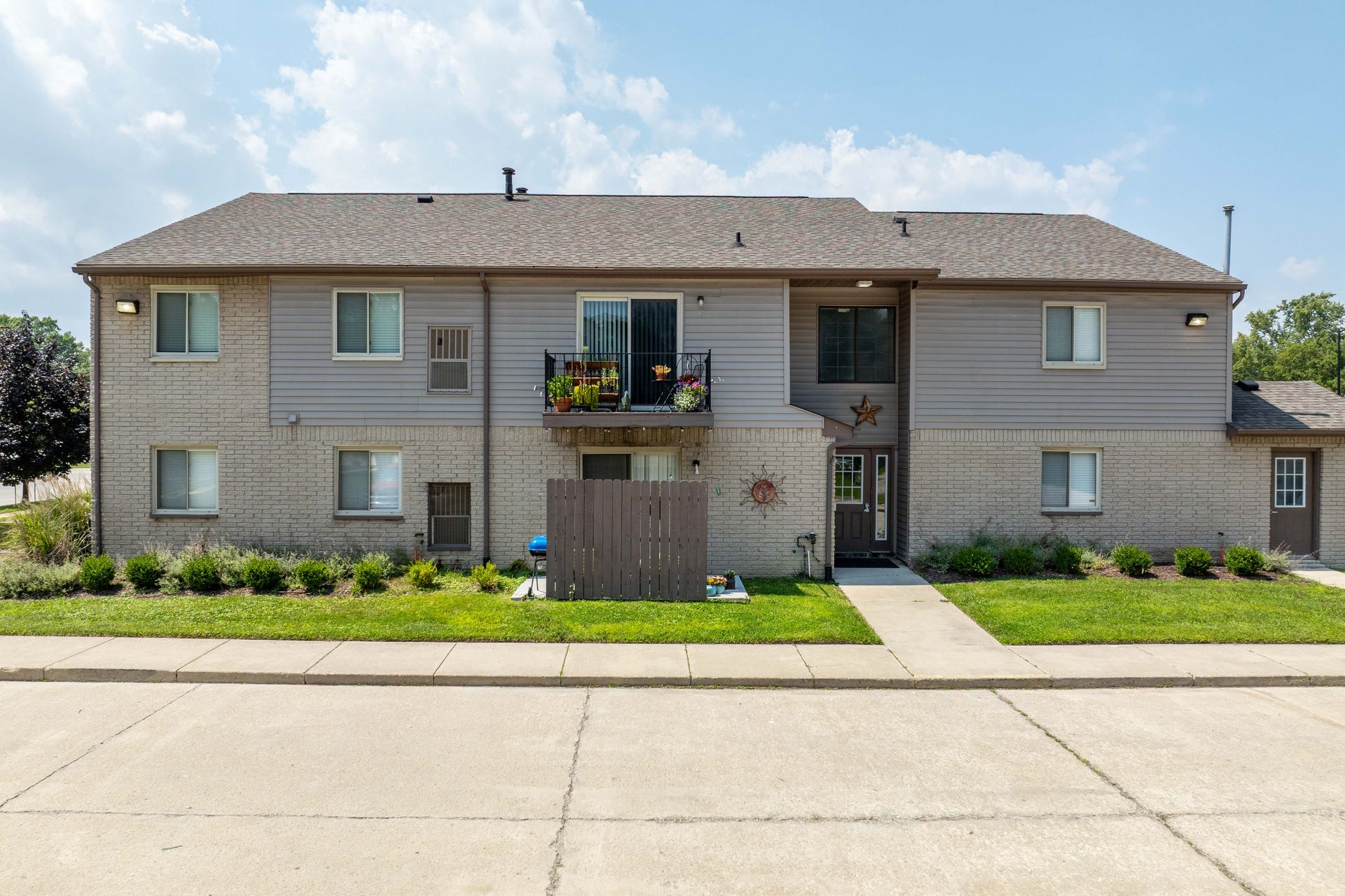 Two-story apartment building with tan and gray exterior, and brown roof. Front view on a sunny day.