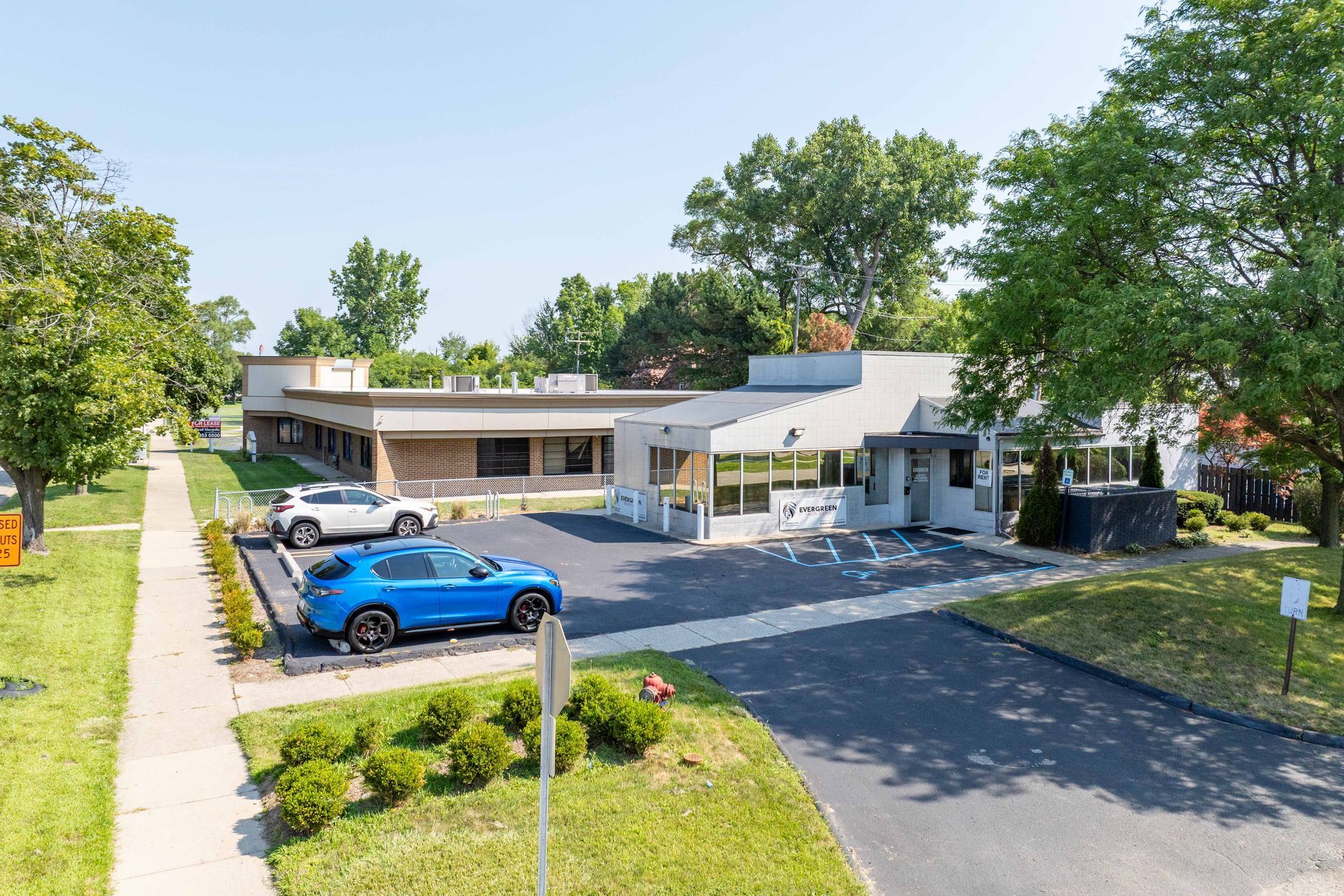 A blue car and a white SUV parked in front of a modern building with a canopy. Green lawn and trees.
