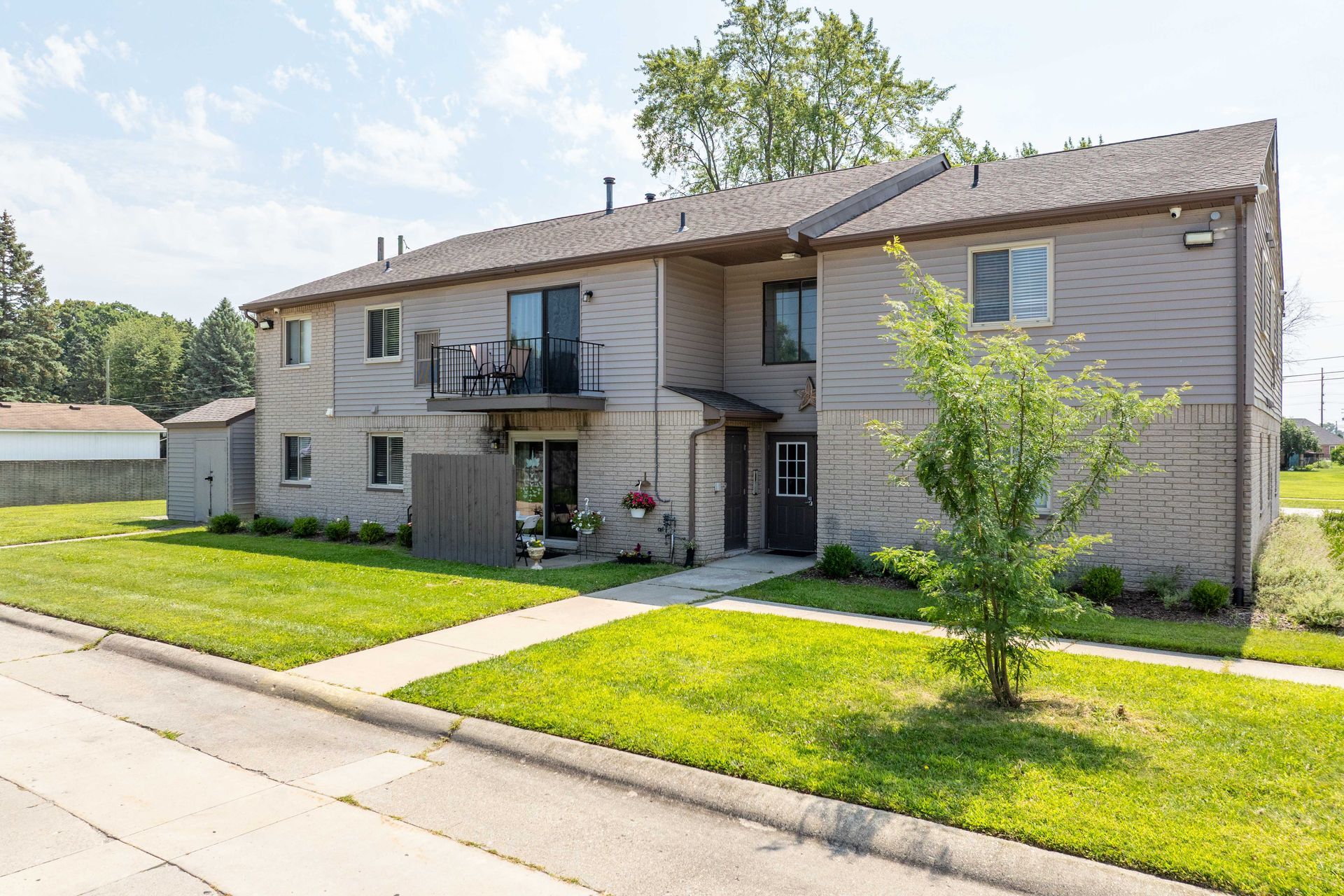 Two-story tan apartment building with green grass and a small tree on a sunny day.