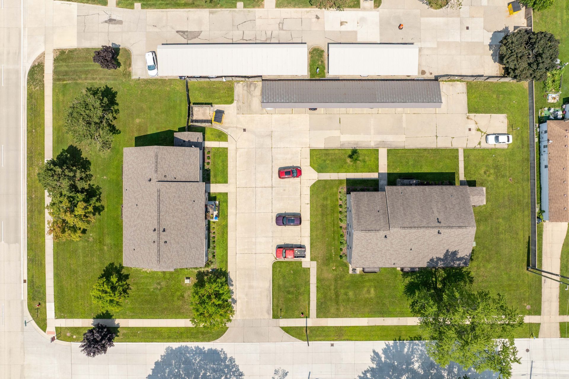Aerial view of a street with two houses, cars parked in between, and a long garage. Green grass and trees are visible.