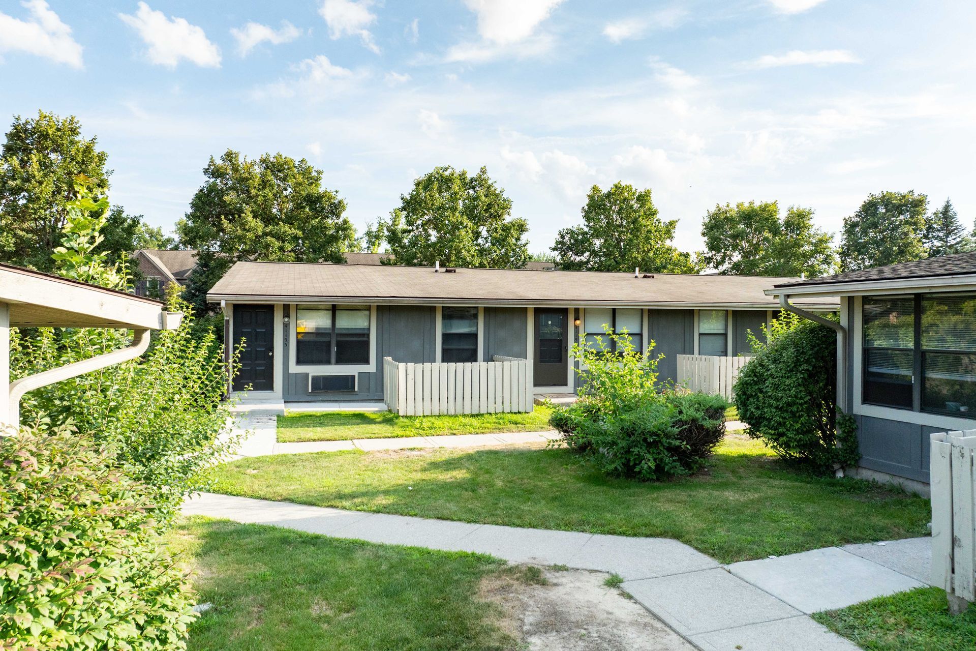 Gray single-story apartments with white picket fences, green lawn, and trees under a blue sky.