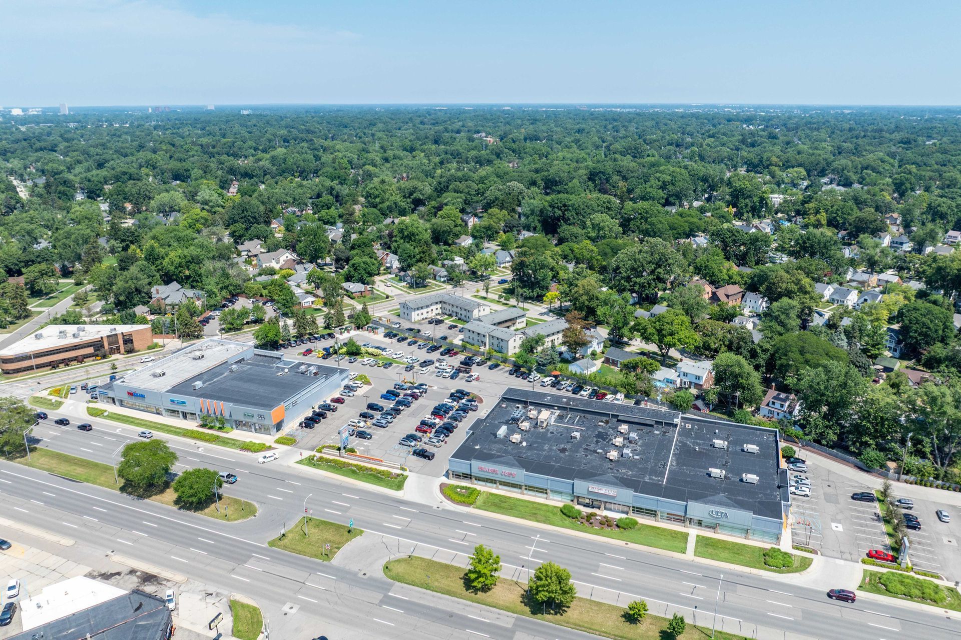 Aerial view of a commercial complex with parking, surrounded by green trees and residential area on a sunny day.
