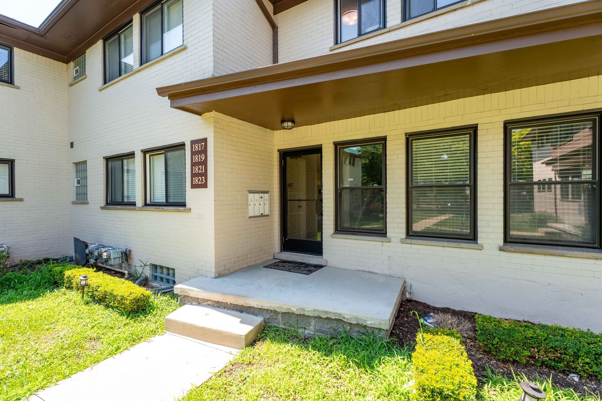 Beige brick apartment building exterior with a covered entrance, a small porch, and a walkway.