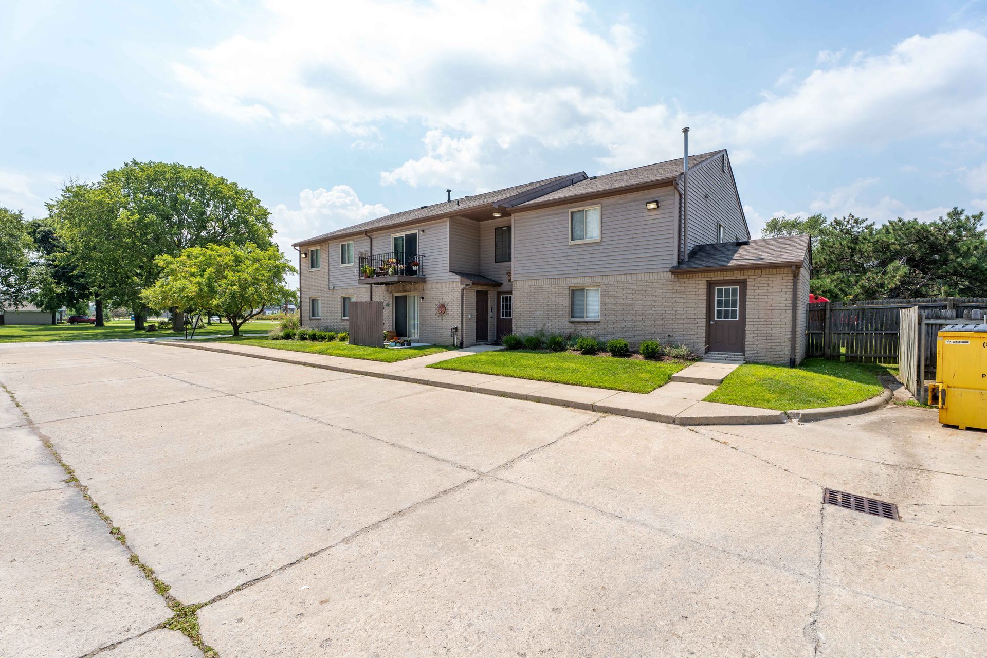 Two-story beige apartment building with a small shed and parking lot on a sunny day.