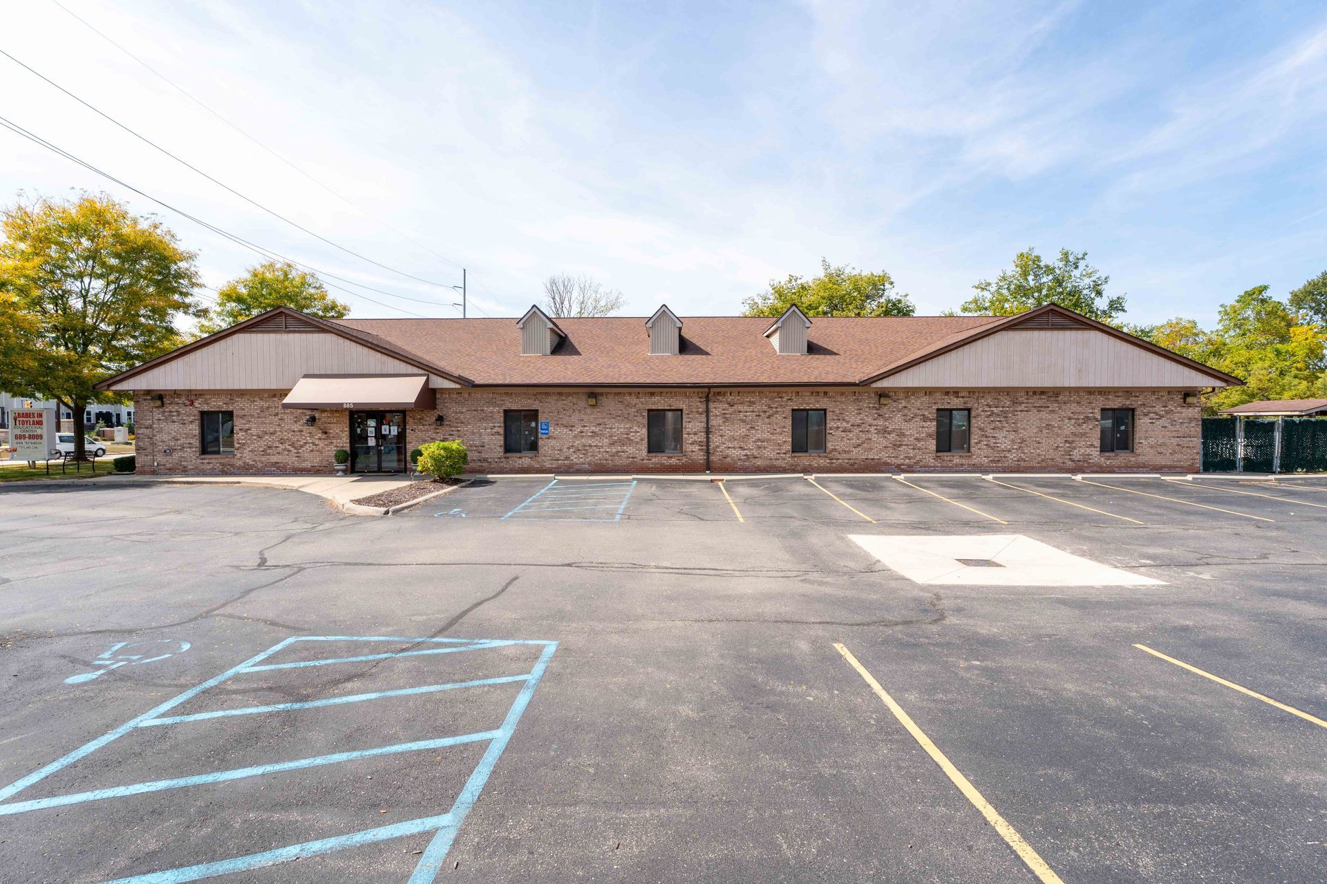 Brick building with brown roof, fronted by a large parking lot. Blue sky visible.