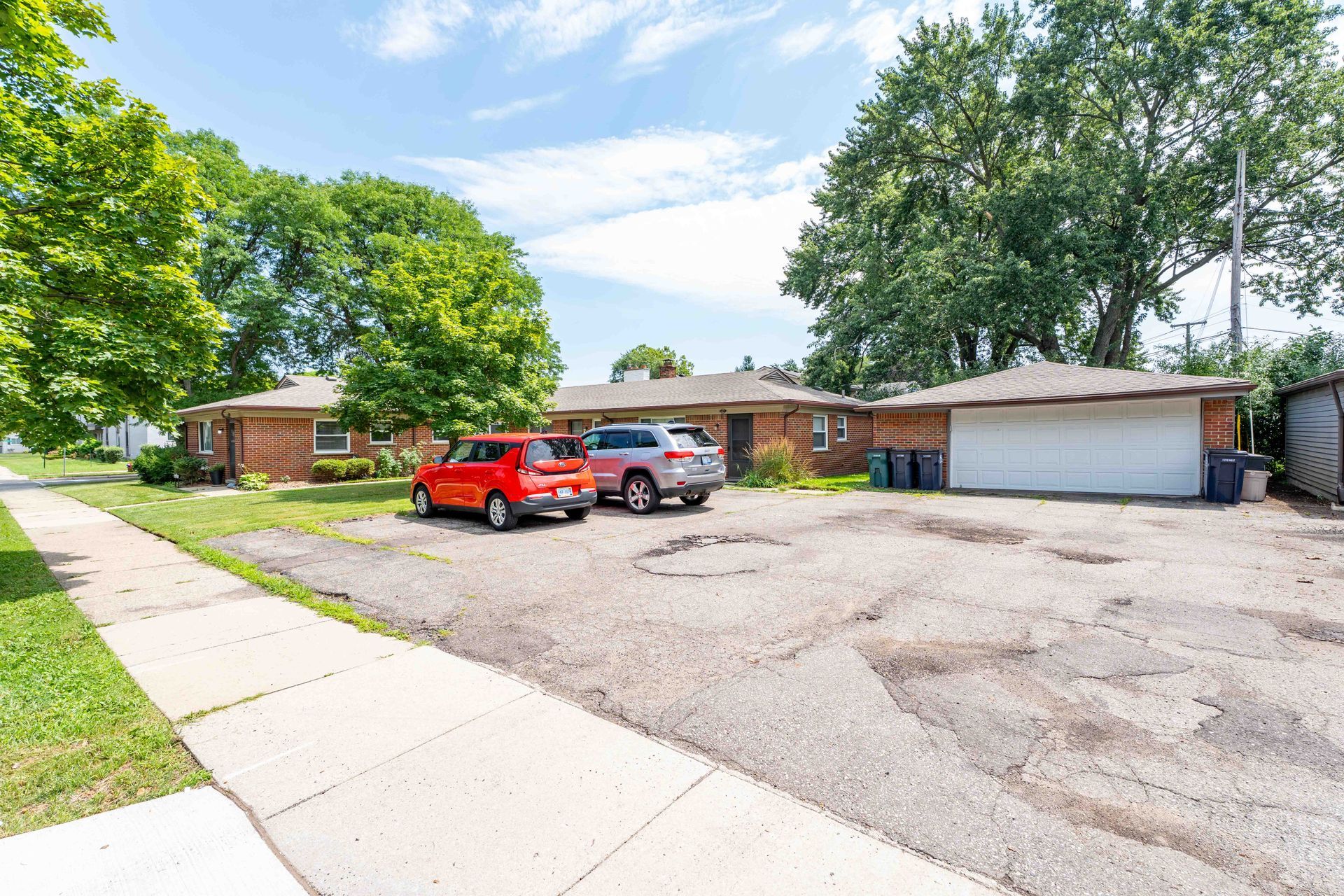 A brick duplex with a two-car garage, vehicles parked in front on a cracked asphalt lot, and a sidewalk.