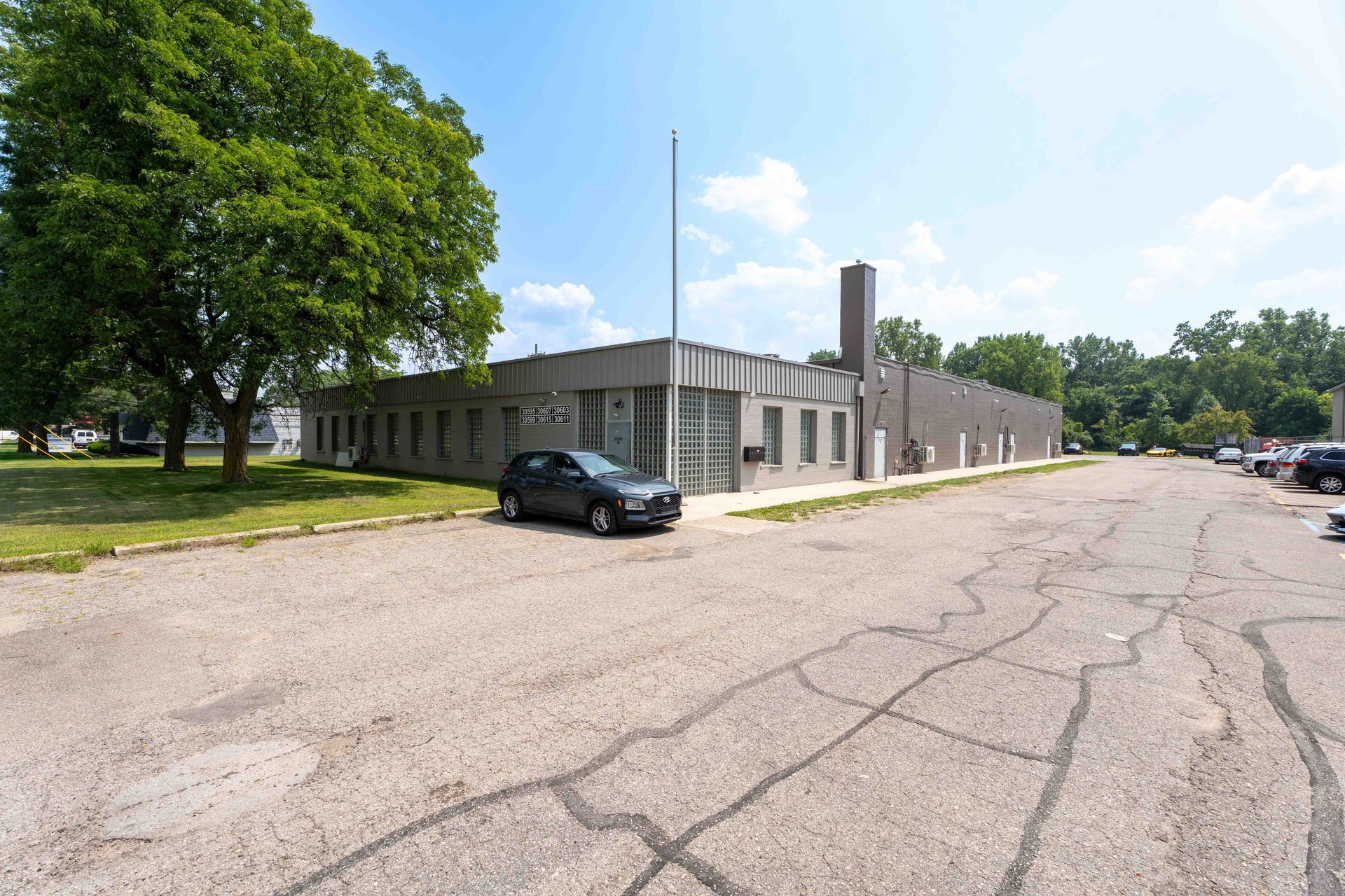 Gray building with a few cars parked in a cracked asphalt lot. Green trees and blue sky visible.
