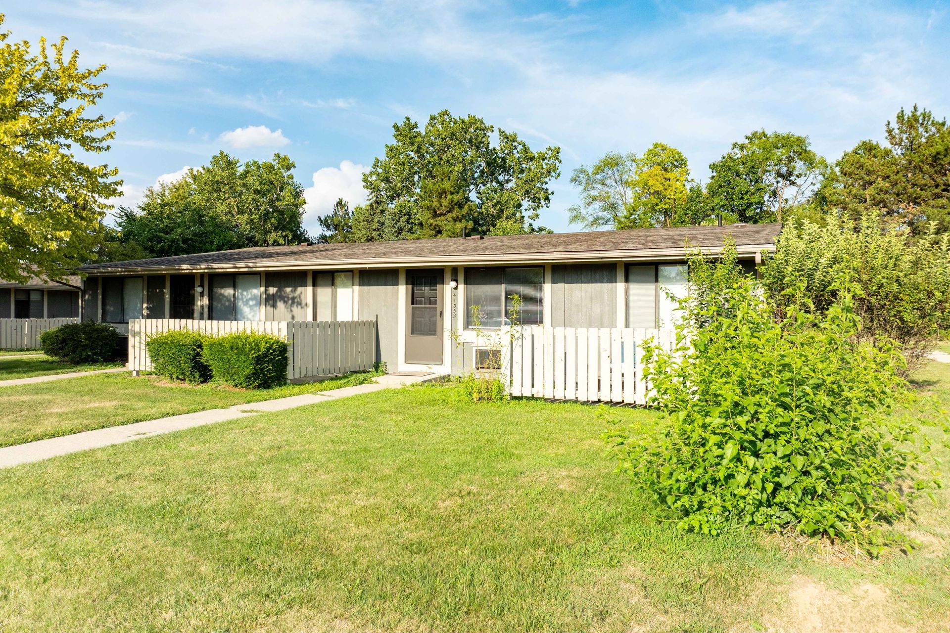 A single-story beige building with a white picket fence, surrounded by green grass and trees under a blue sky.