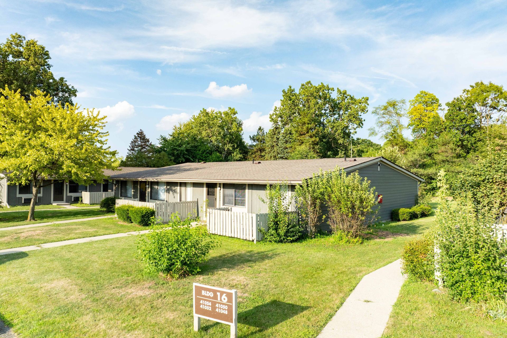 Exterior of a single-story apartment building with green grass, trees, and a blue sky.