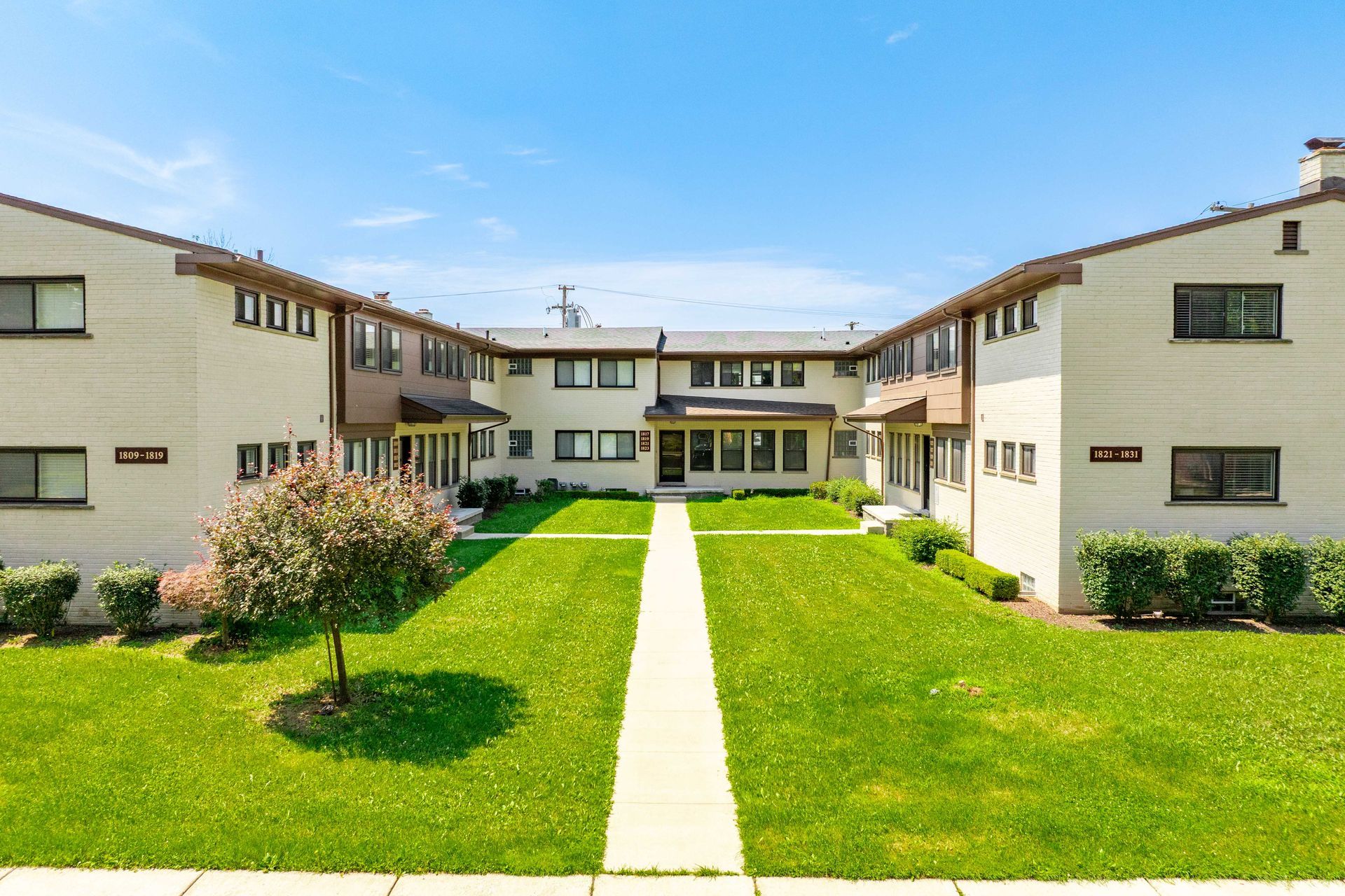 Low-rise apartment complex with symmetrical lawn, light beige buildings, and bright blue sky.