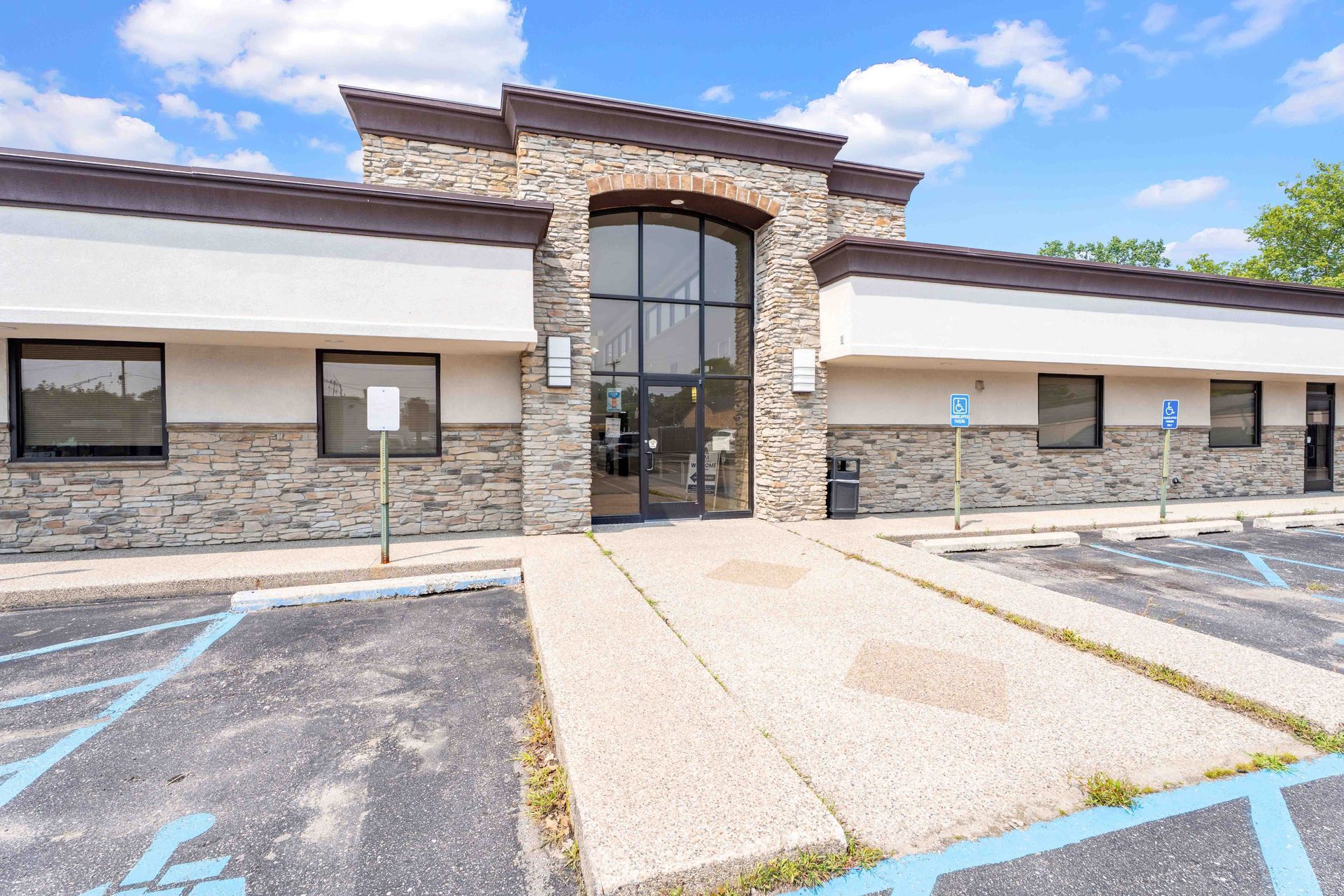 Exterior of a light tan building with stone accents, featuring accessible ramp, parking, and large glass door entrance.