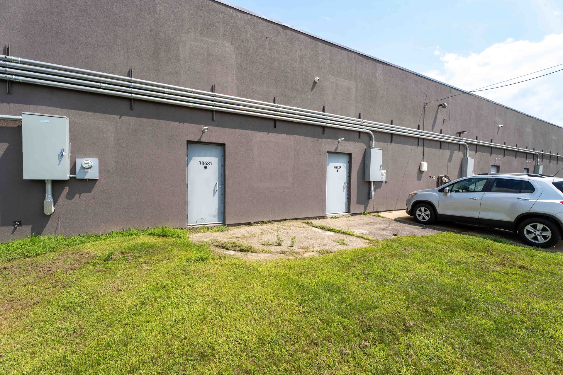 Exterior of a gray building with two doors, electrical boxes, and a car parked nearby. Green grass in foreground.