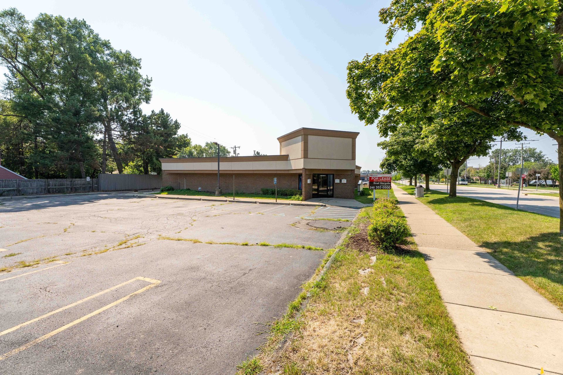 A vacant commercial building with a large empty parking lot and sidewalk alongside a street with trees.
