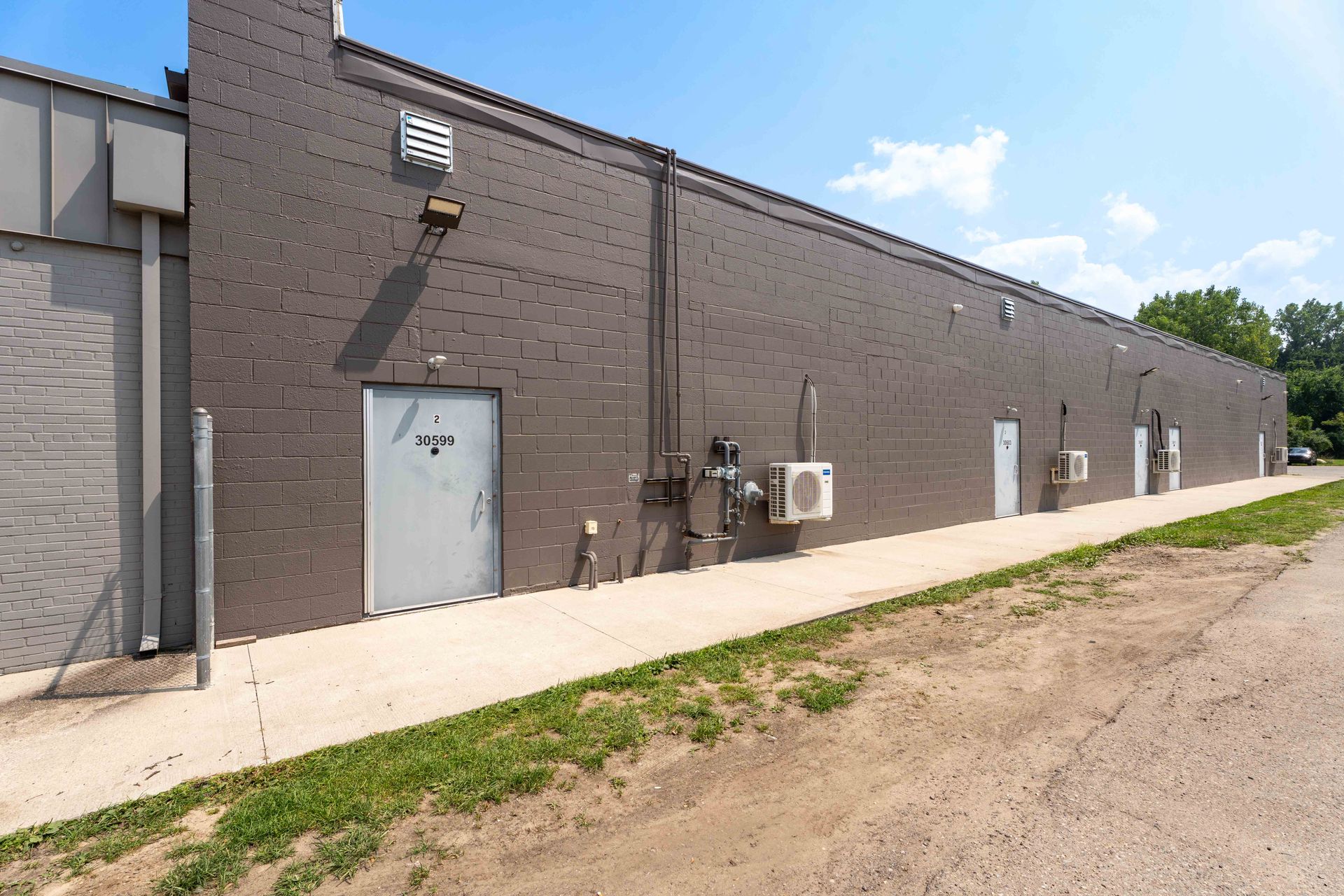 Gray brick building with several white doors, air conditioning units, and a sidewalk.