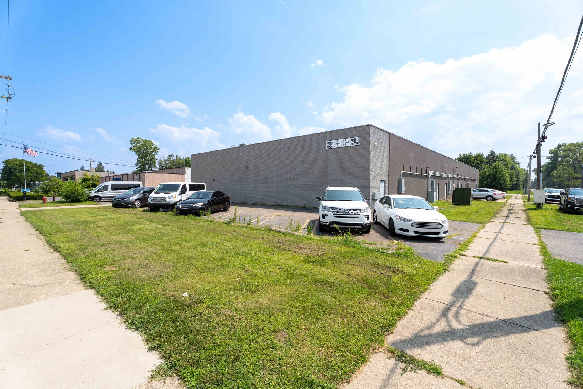Exterior of a gray commercial building with several cars parked on a grassy lot next to a sidewalk.
