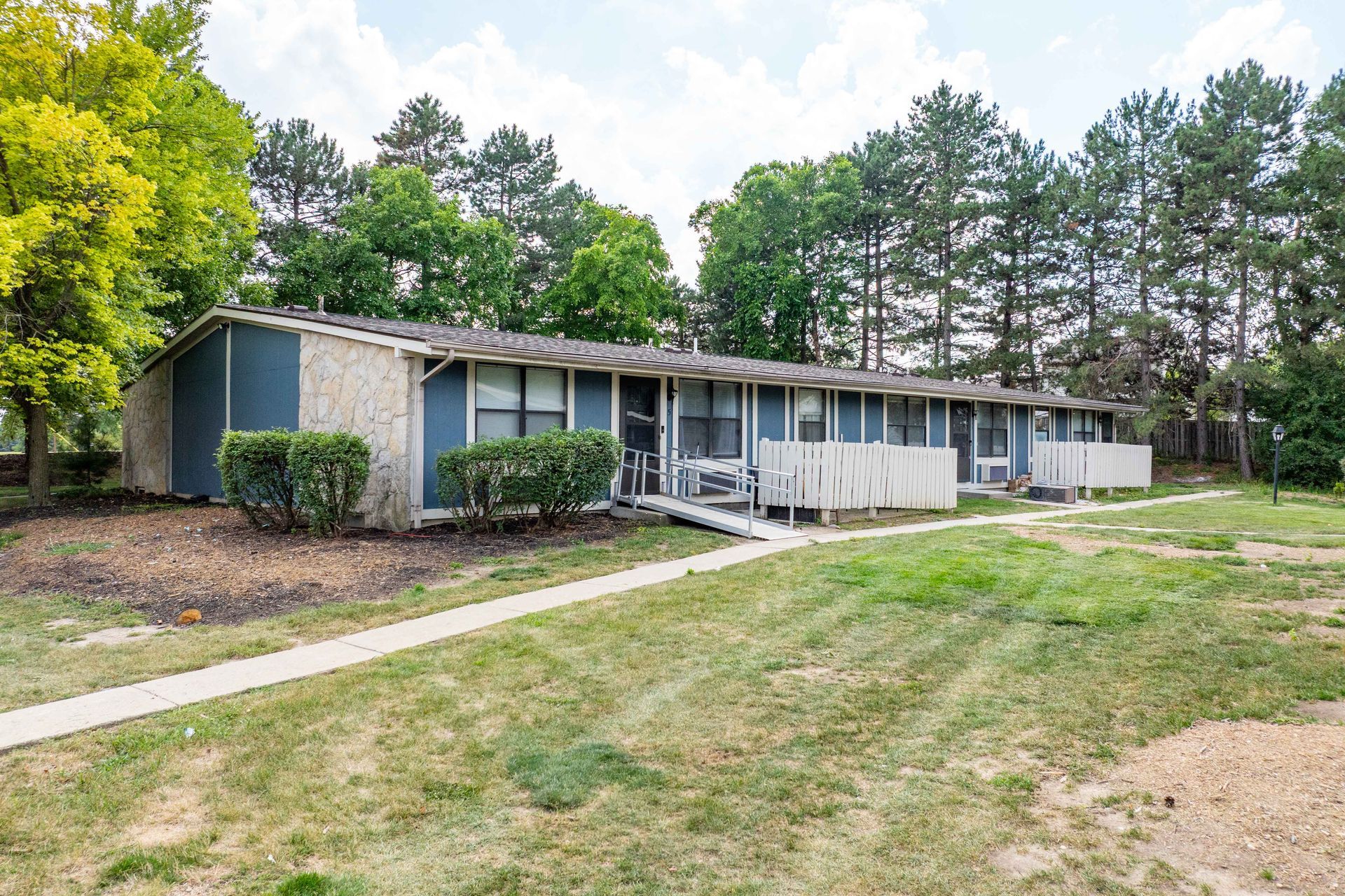 Low-rise apartment building with teal siding, off-white fencing, and a concrete path leading to the doors.