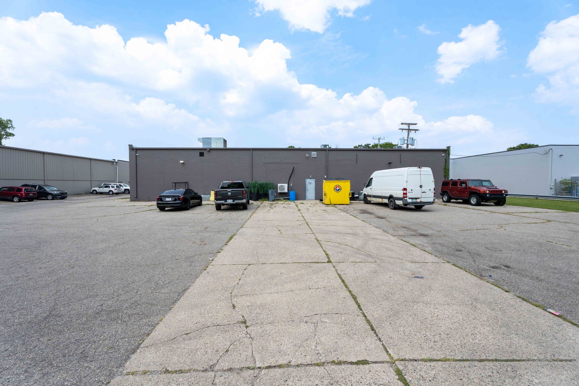 Cars parked in a paved lot in front of a gray industrial building on a partly cloudy day.