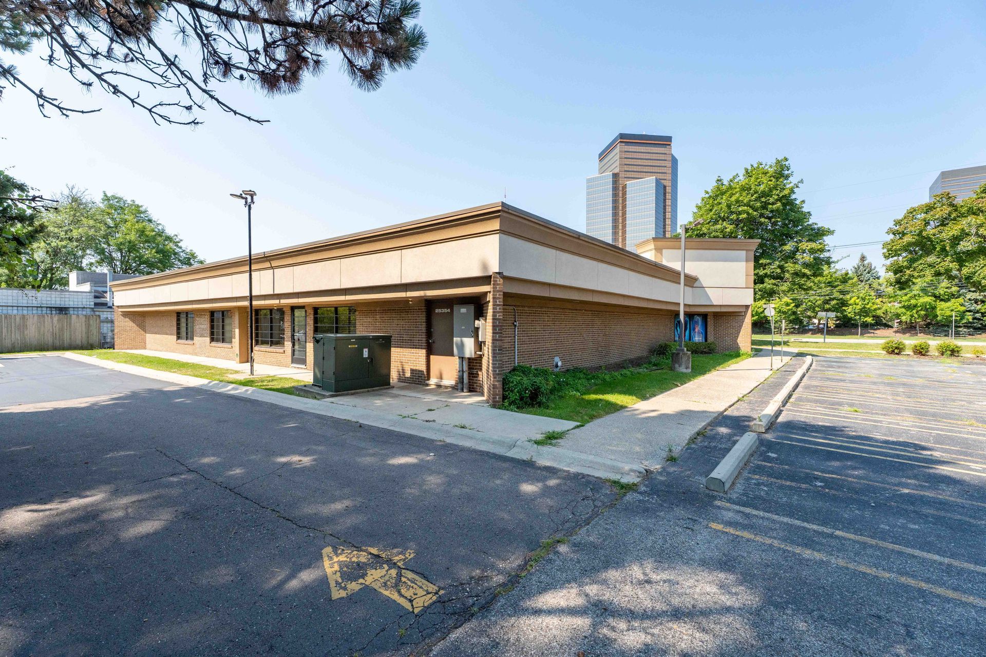 Low-slung, tan brick commercial building with a paved parking lot; trees and a tall building in the background.