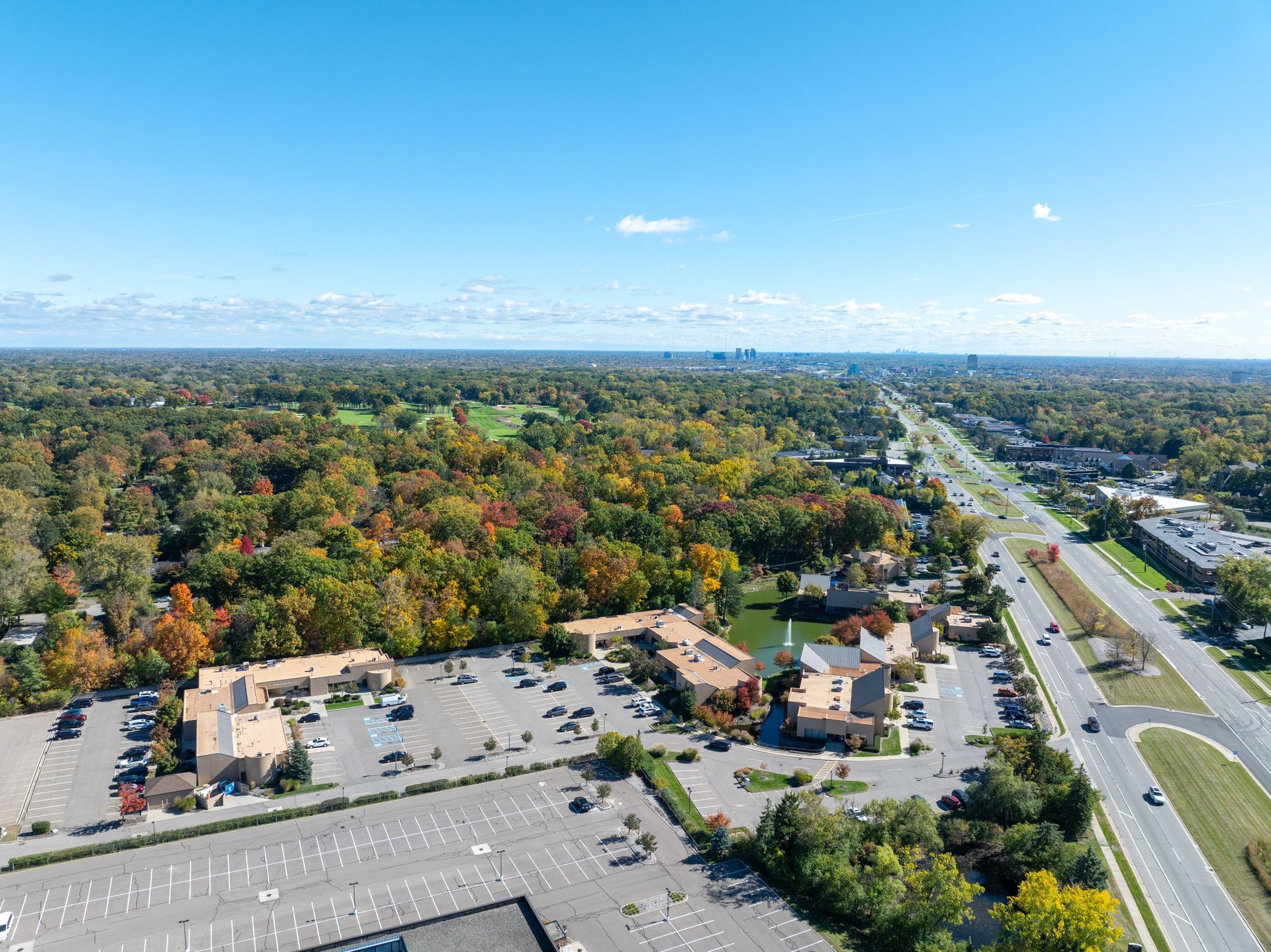 Aerial view of a commercial area with buildings, parking, and a highway surrounded by colorful trees under a blue sky.