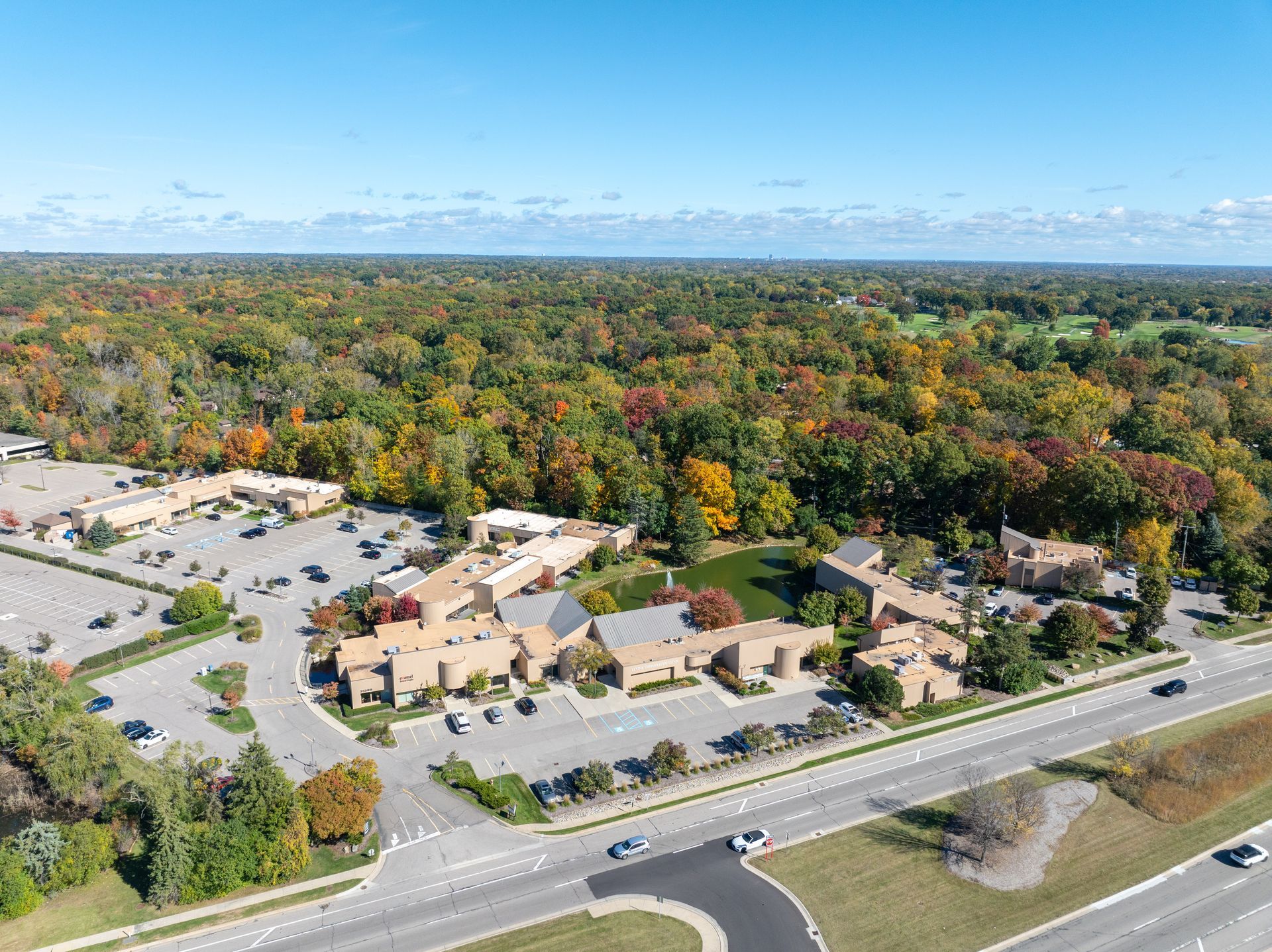 Aerial view of a shopping center with brown buildings, a parking lot, and a dense forest with colorful fall foliage.