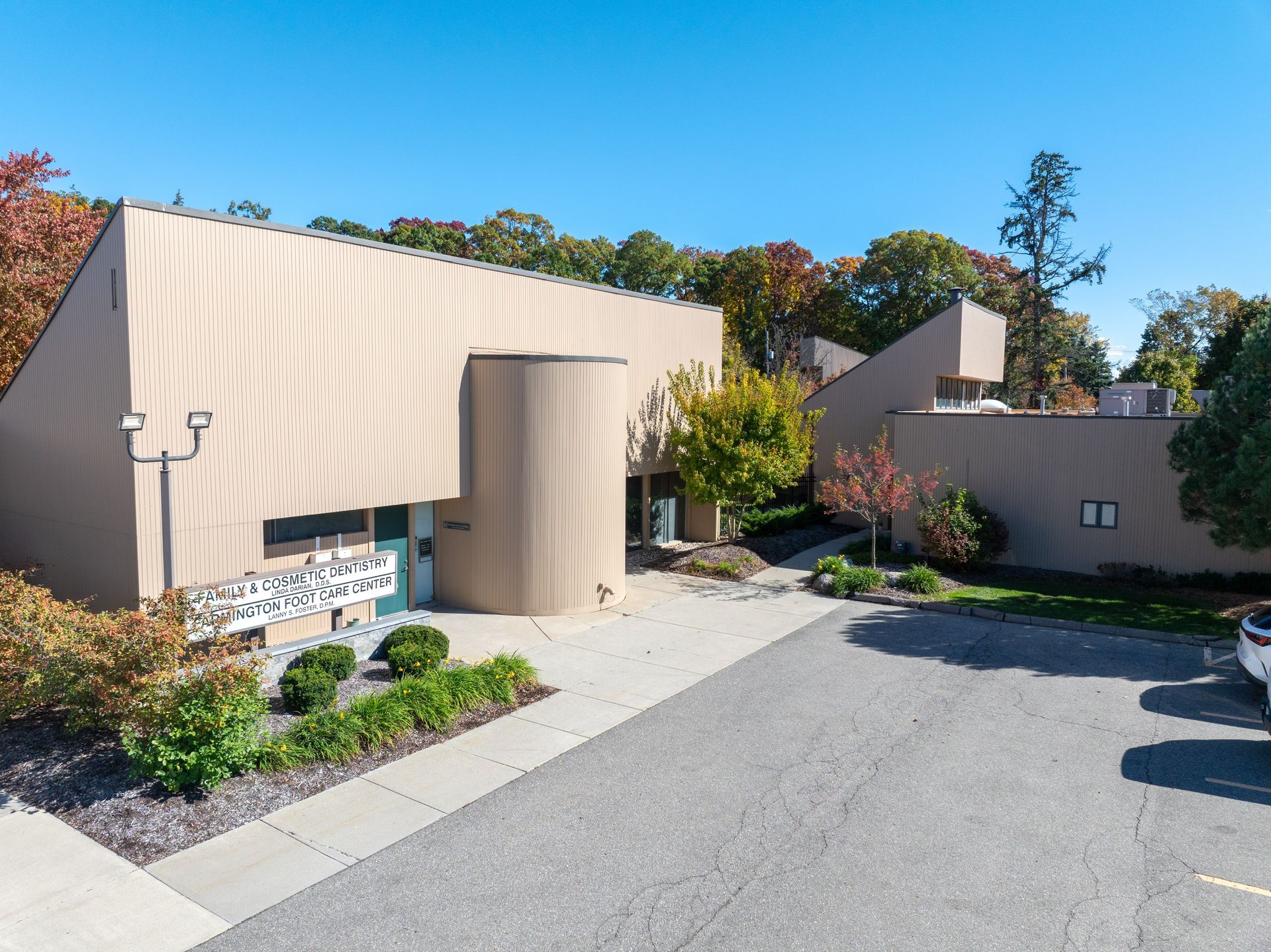 Modern tan building with curved entryway and small parking area, surrounded by trees.