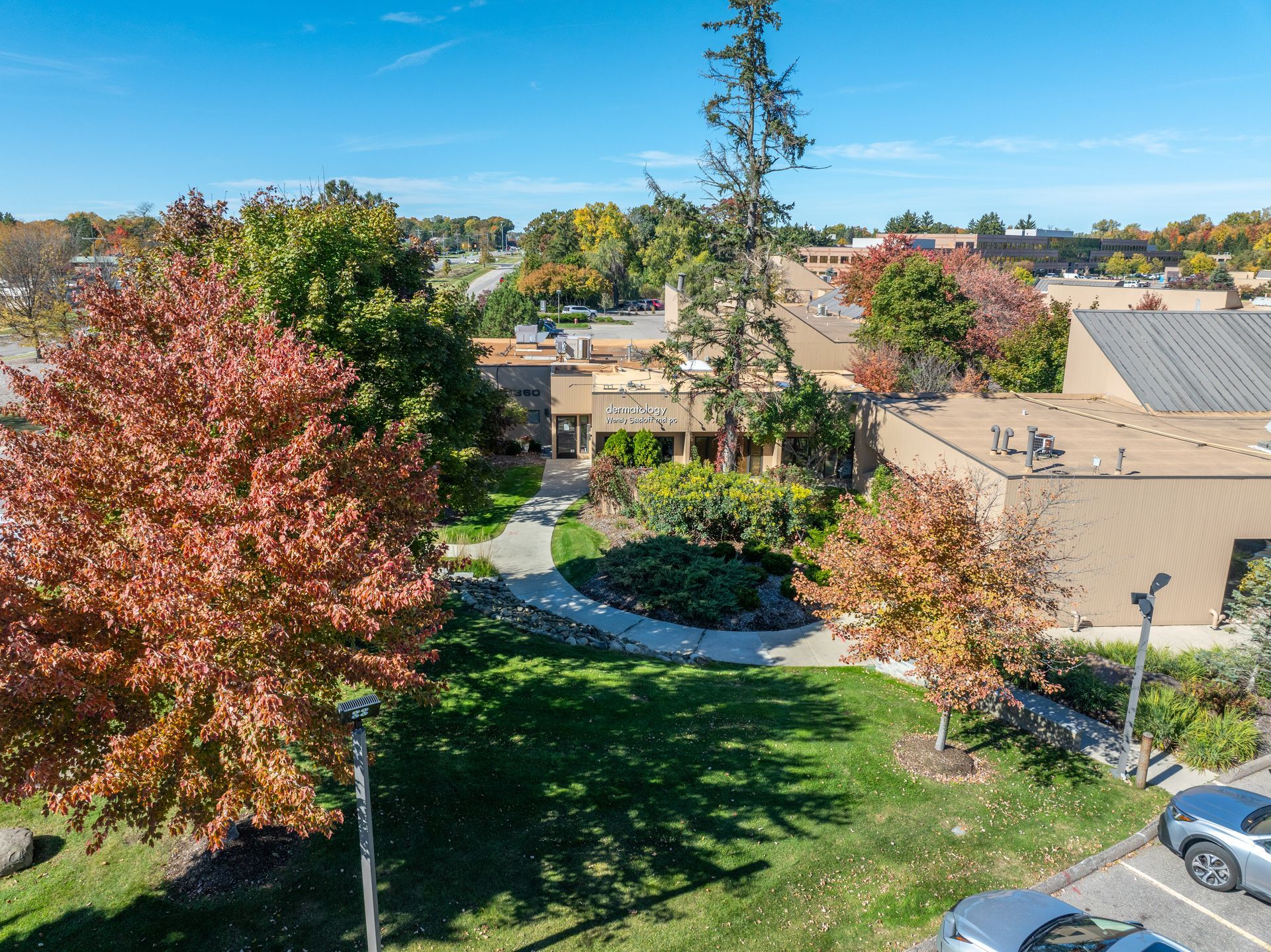 An aerial view of a building and surrounding green space with trees showing fall colors and parked cars.