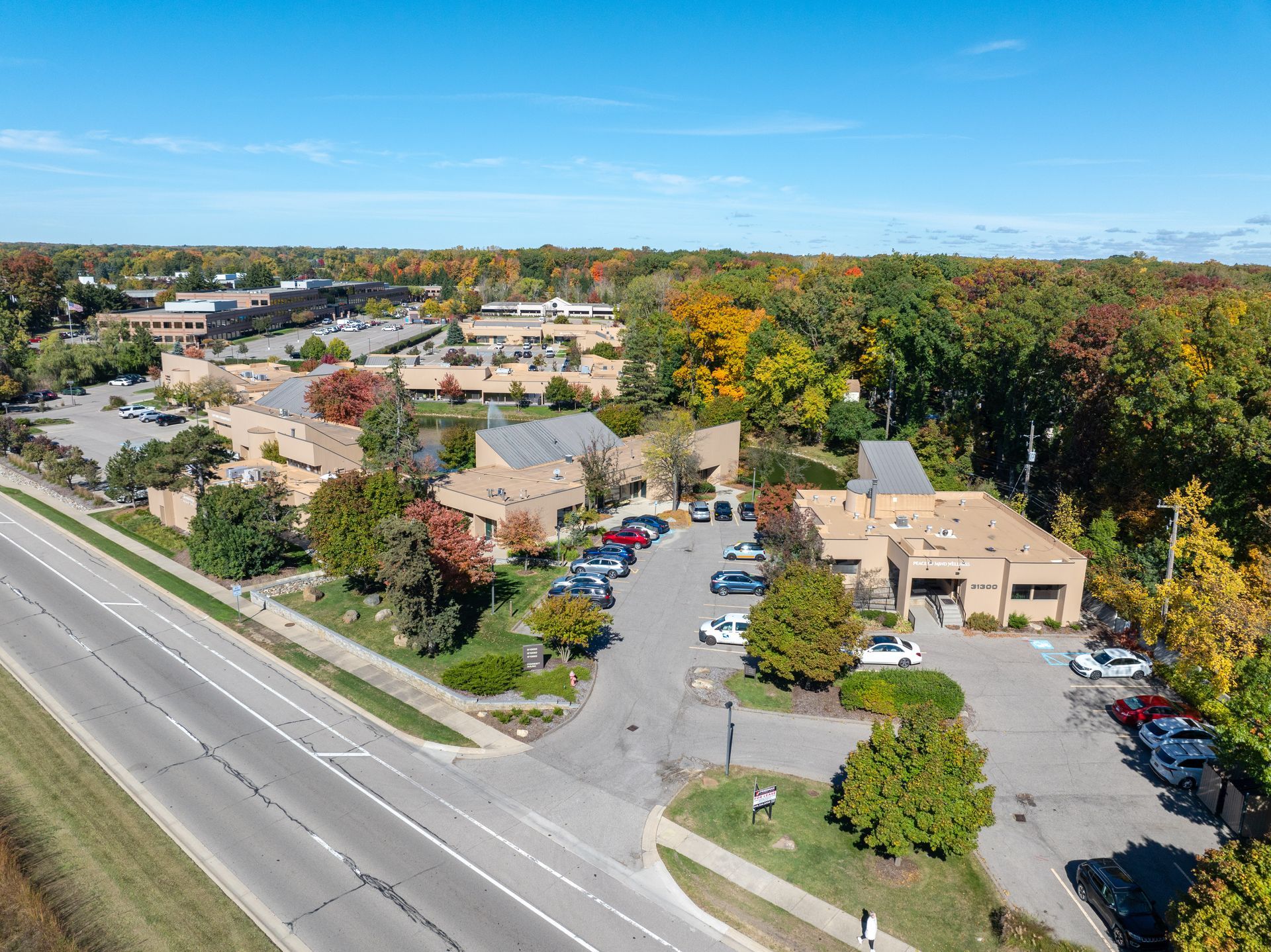 Aerial view of a commercial complex next to a highway with parked cars and surrounding autumn trees under a blue sky.