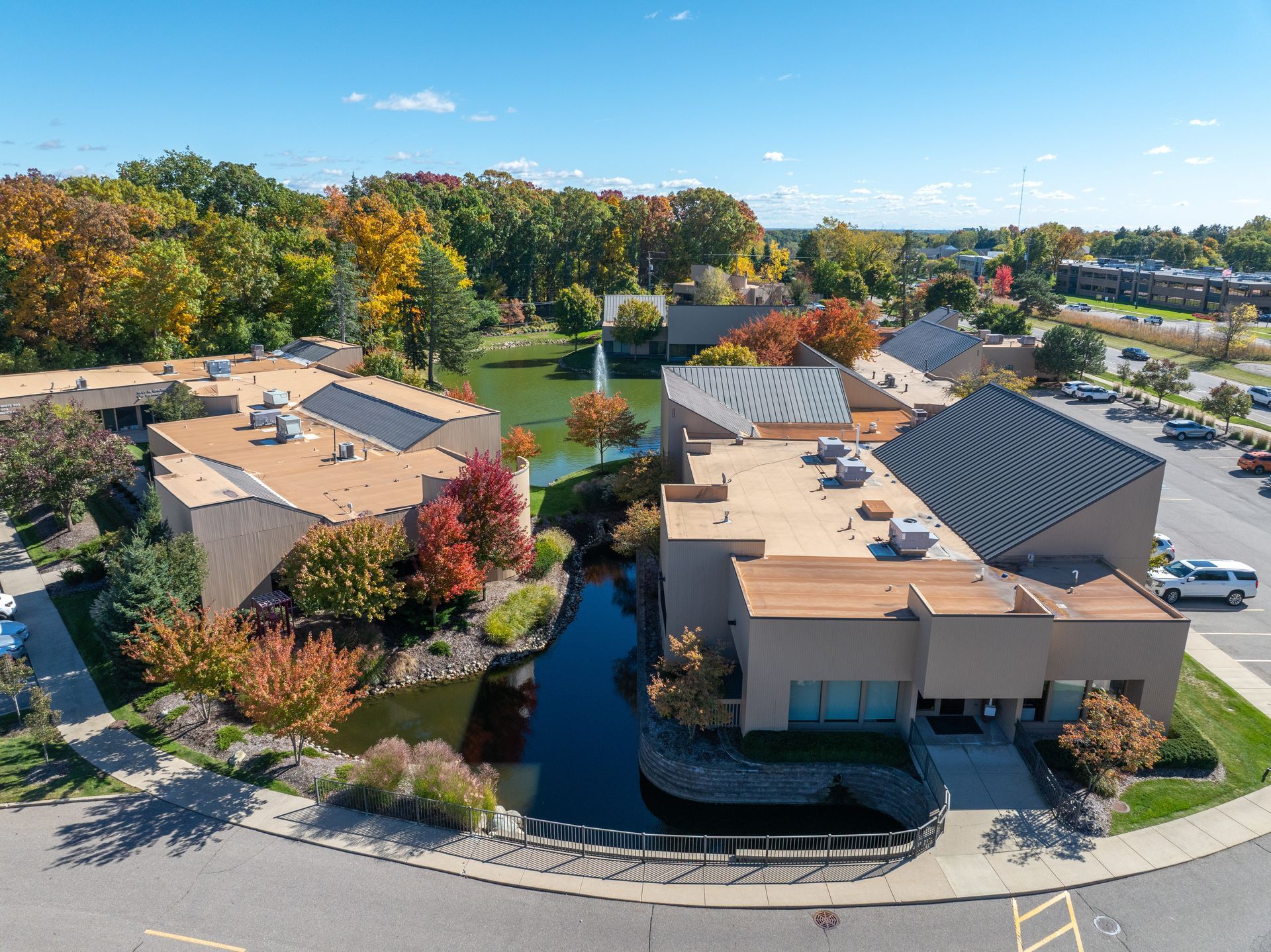 Aerial view of a complex with buildings, a pond, and trees in autumn. A parking lot is to the right.