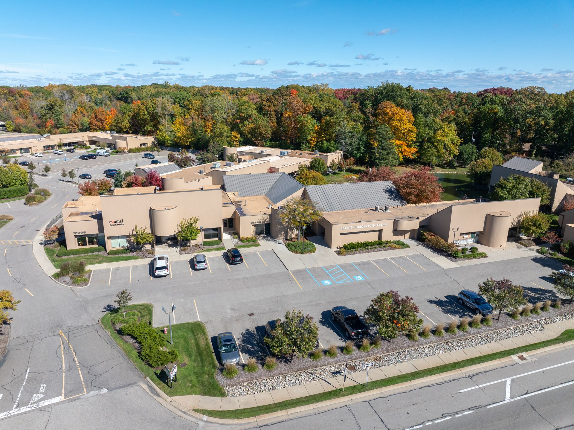 An aerial view of a beige building with a parking lot and trees; sunny day.